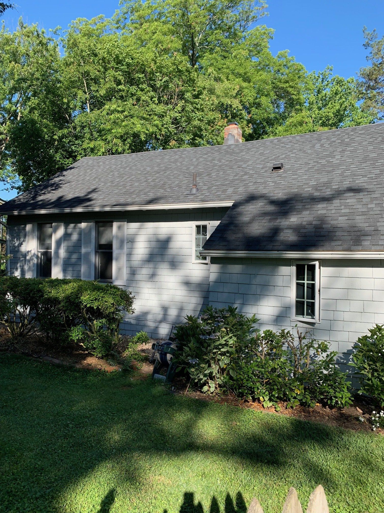 Single-story gray house with a shingled roof, surrounded by green lawn and trees in sunlight.