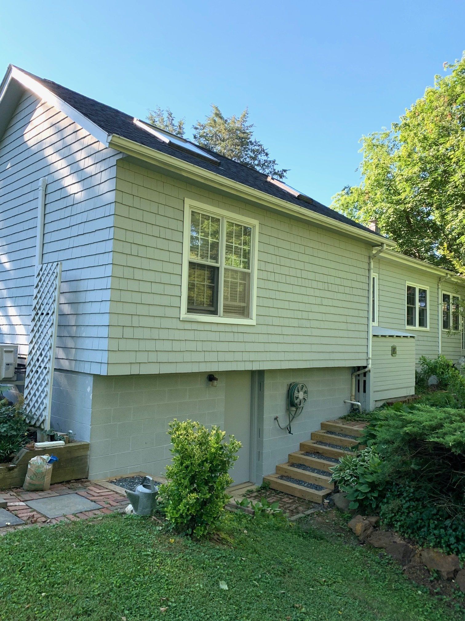 Side view of a light-colored house with a sloped roof, windows, stairs, and a grassy yard with trees.