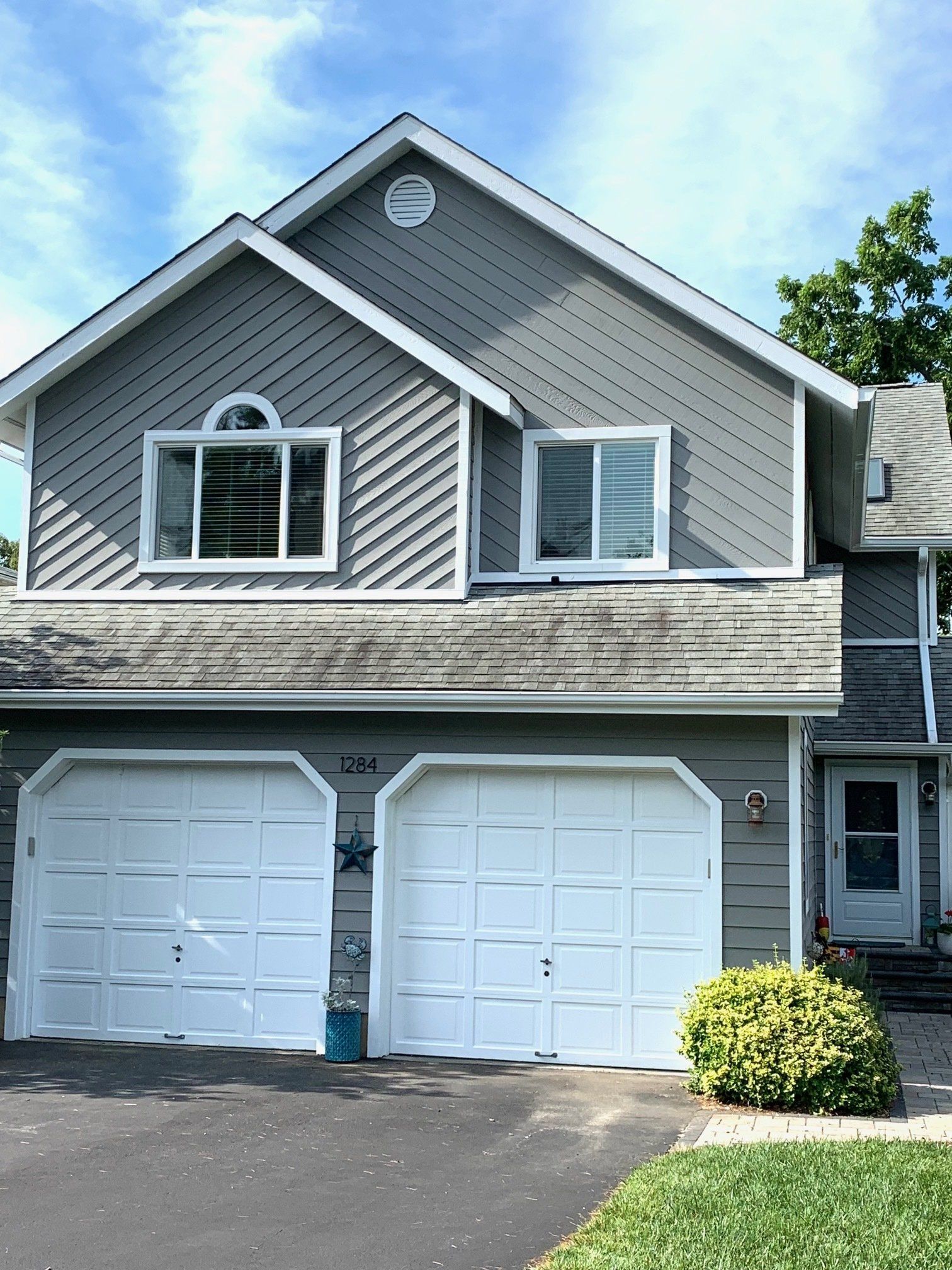 Two-story gray house with white double garage doors, dormer windows, and a paved driveway.