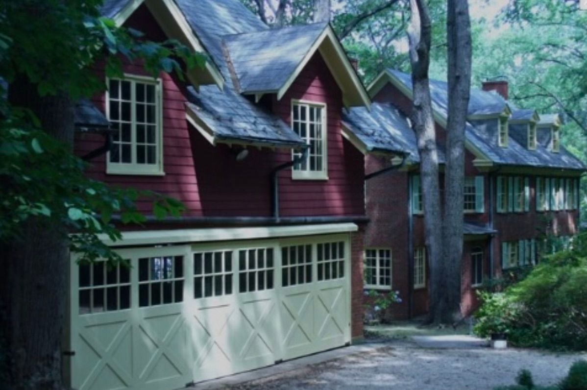 Red house with white garage doors and dormer windows on a wooded driveway
