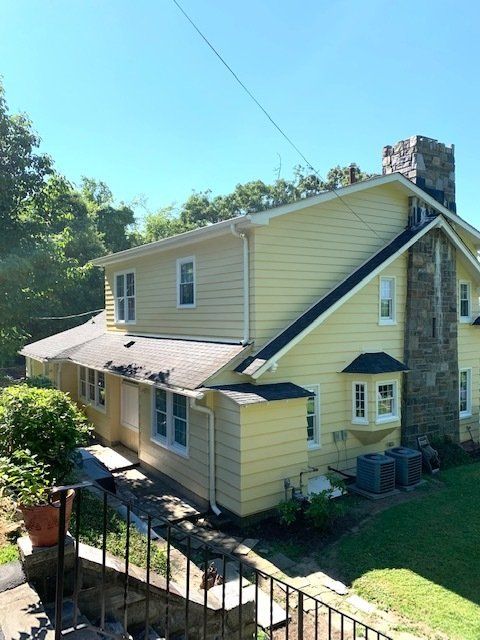 Yellow two-story house with stone chimney, white trim, and a sloped roof on a wooded hillside