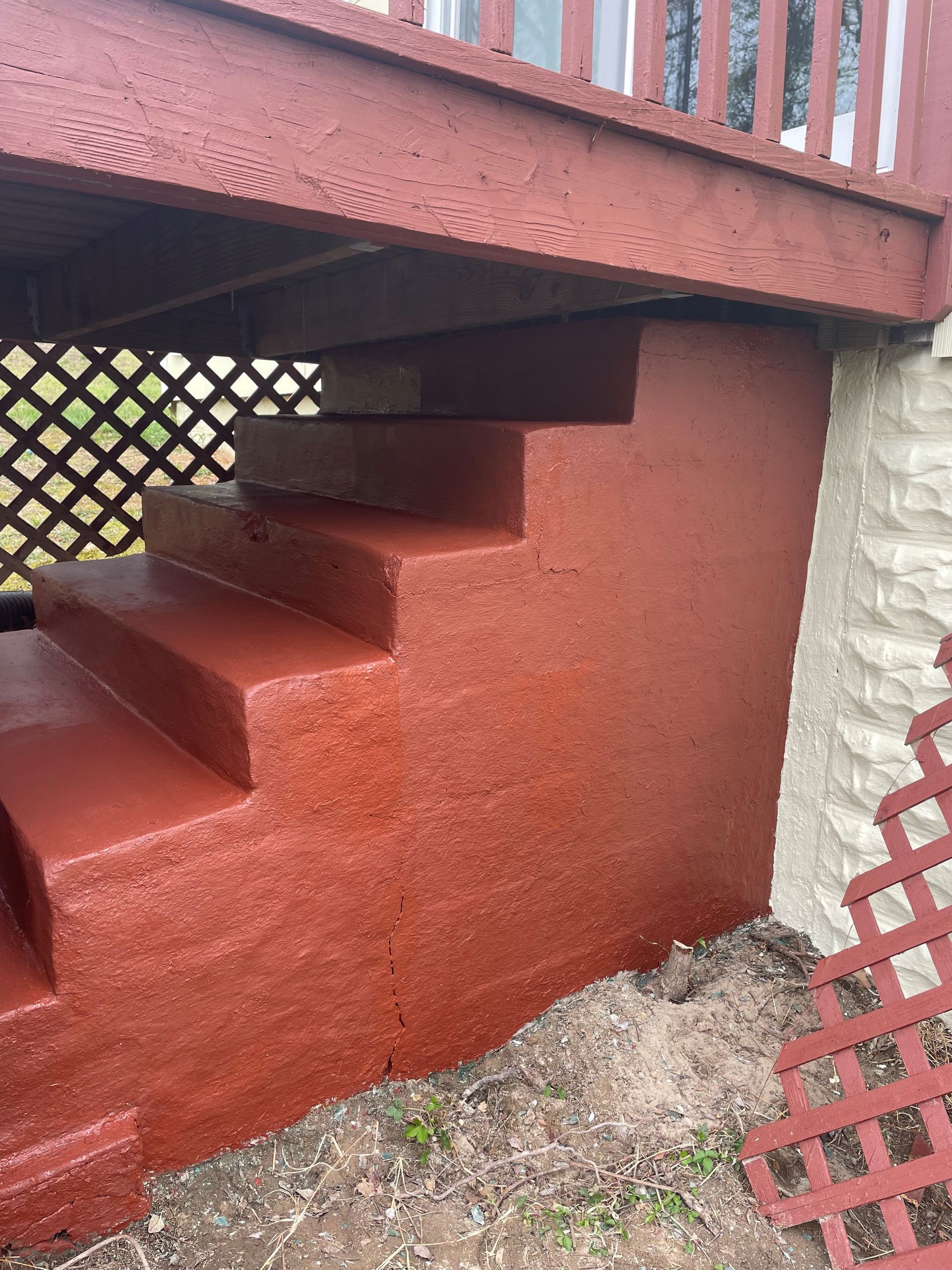 Red-painted exterior stairs under a wooden deck beside a lattice fence and stone wall.