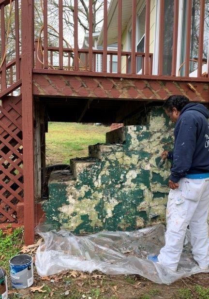 Person painting the stone foundation of a deck with green and white camouflage paint.