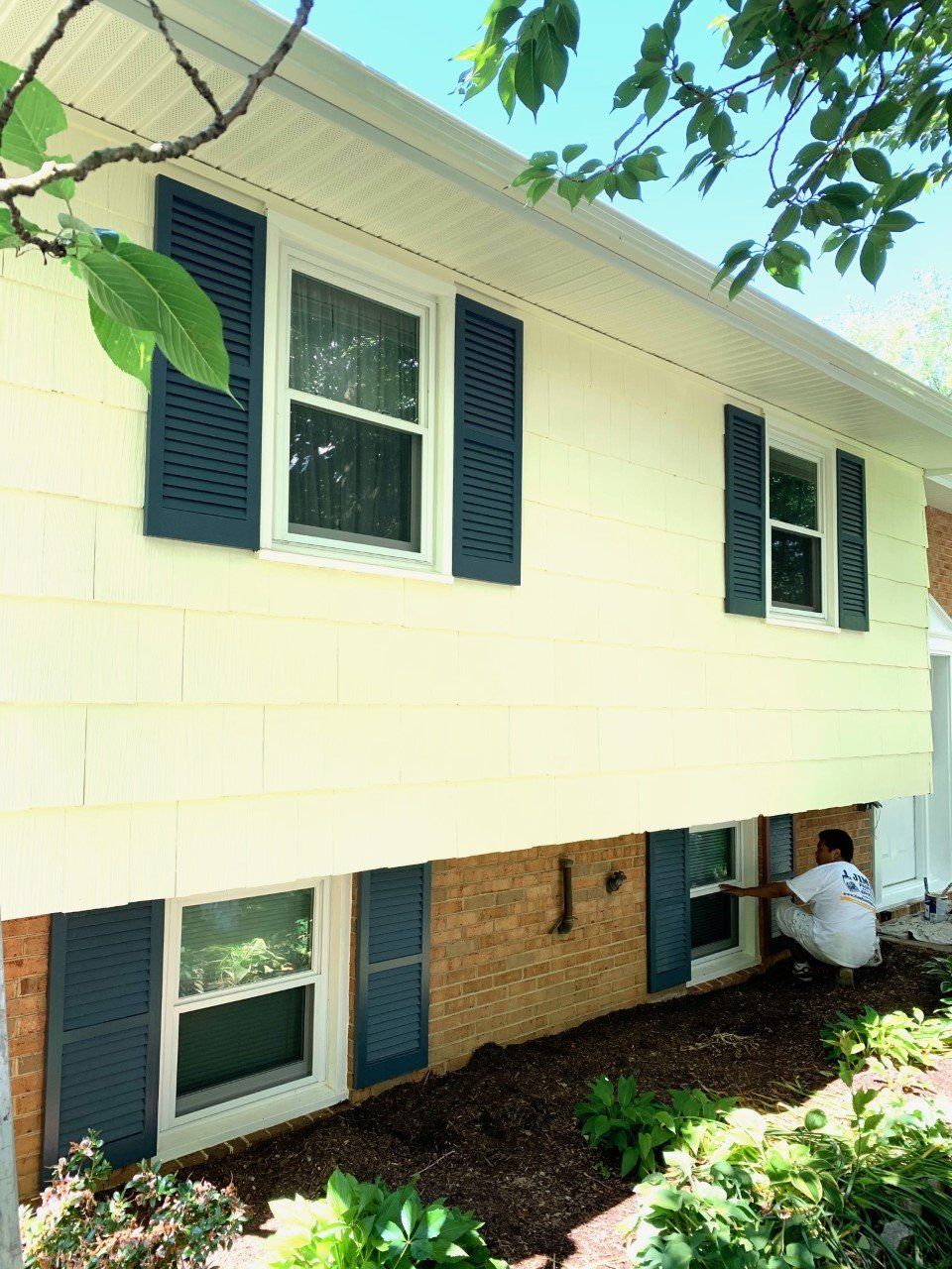 Raised house exterior with white siding, blue shutters, and brick lower level, seen from a leafy yard