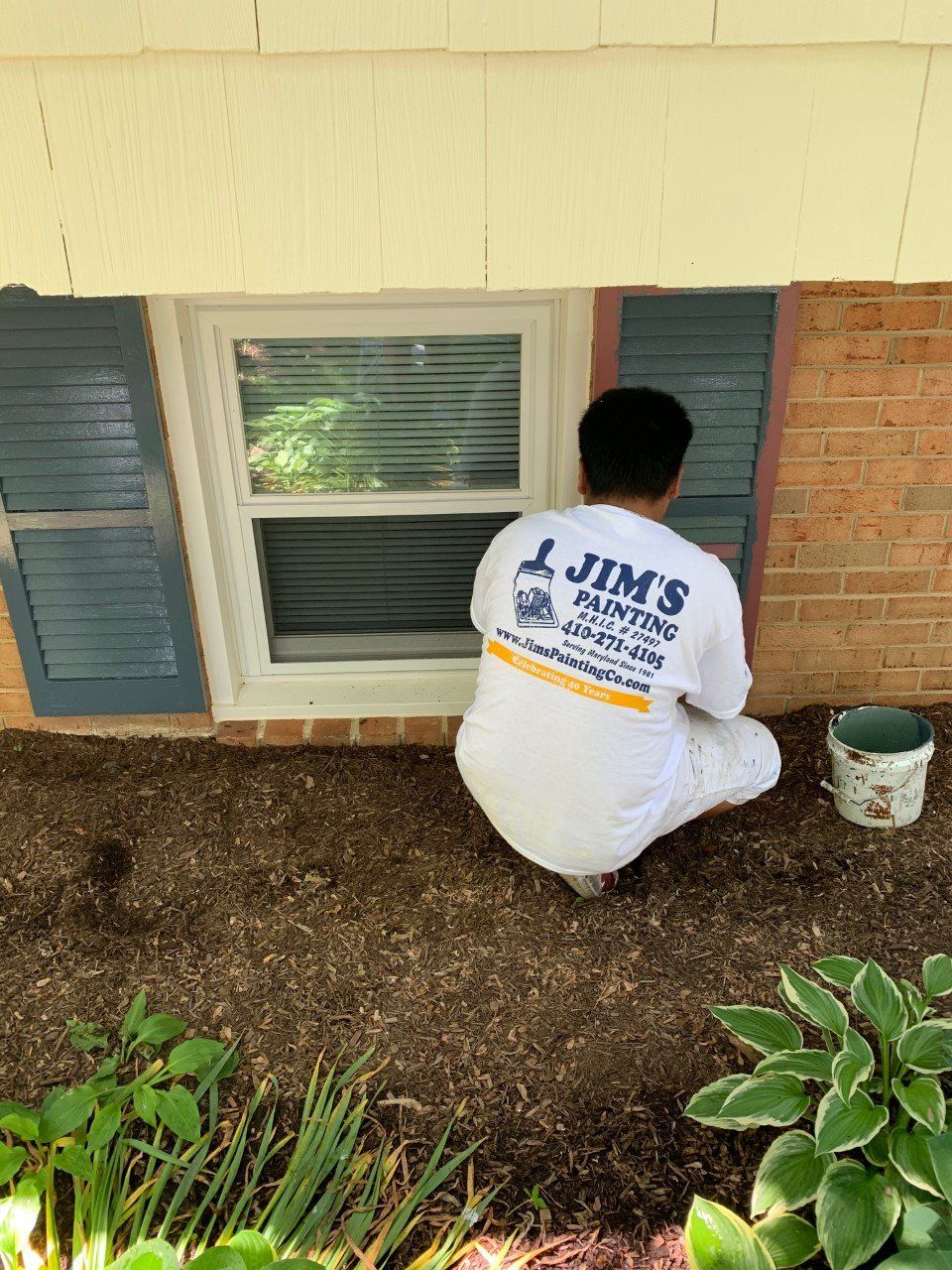 Person kneeling by a house window, painting the exterior wall near blue shutters and a white bucket.