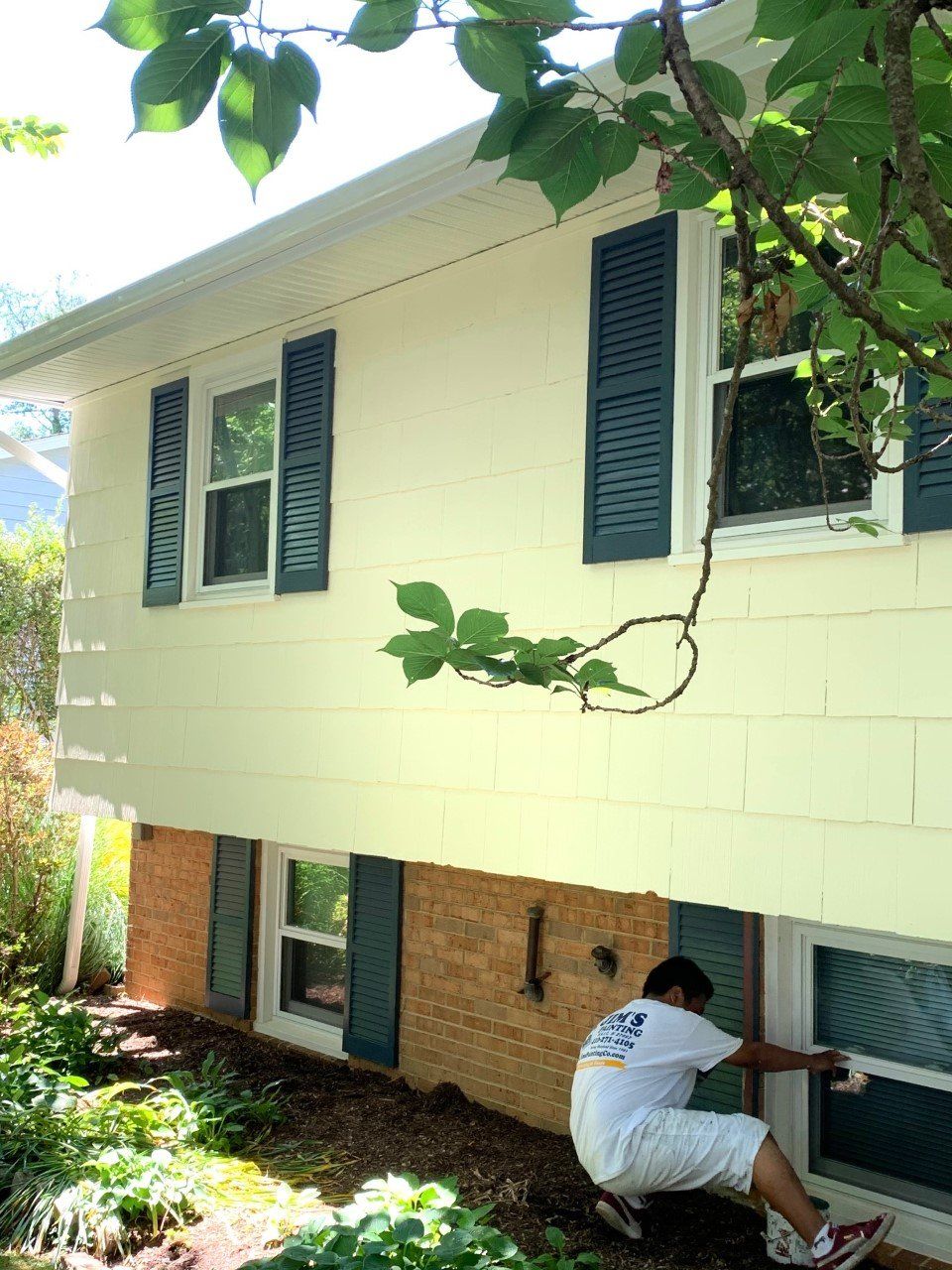 Person crouching under a raised house, painting the lower exterior wall and trim.