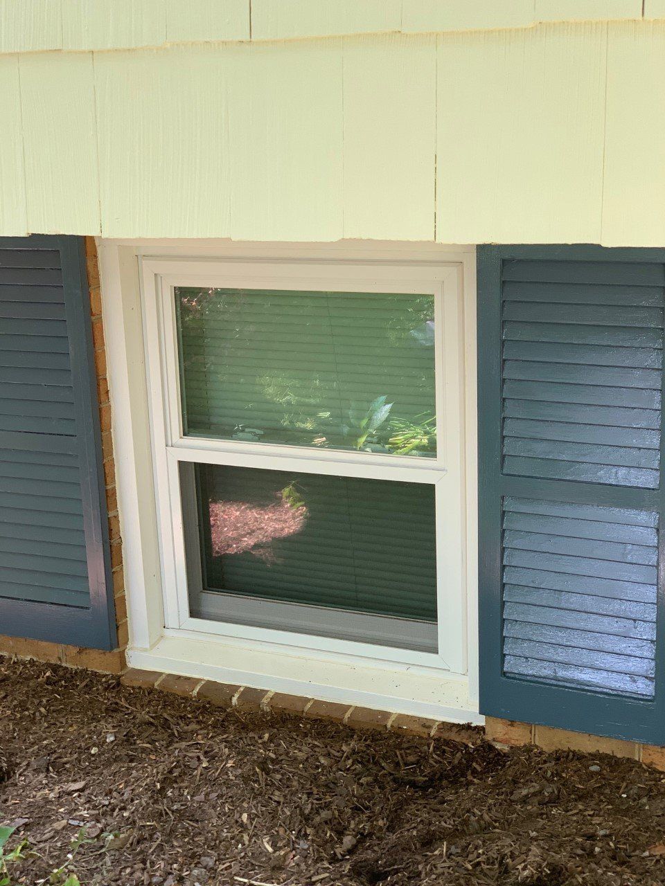 White house window with blue shutters and partially open blinds, set in a mulch bed outside.
