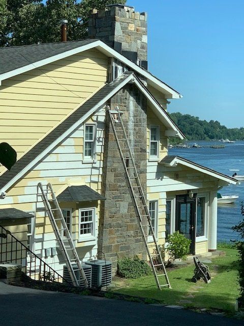 Stone and yellow lakeside house with a steep roof, ladder, and water view on a sunny day