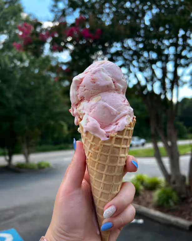 Hand holding a waffle cone with two scoops of pale pink ice cream outdoors