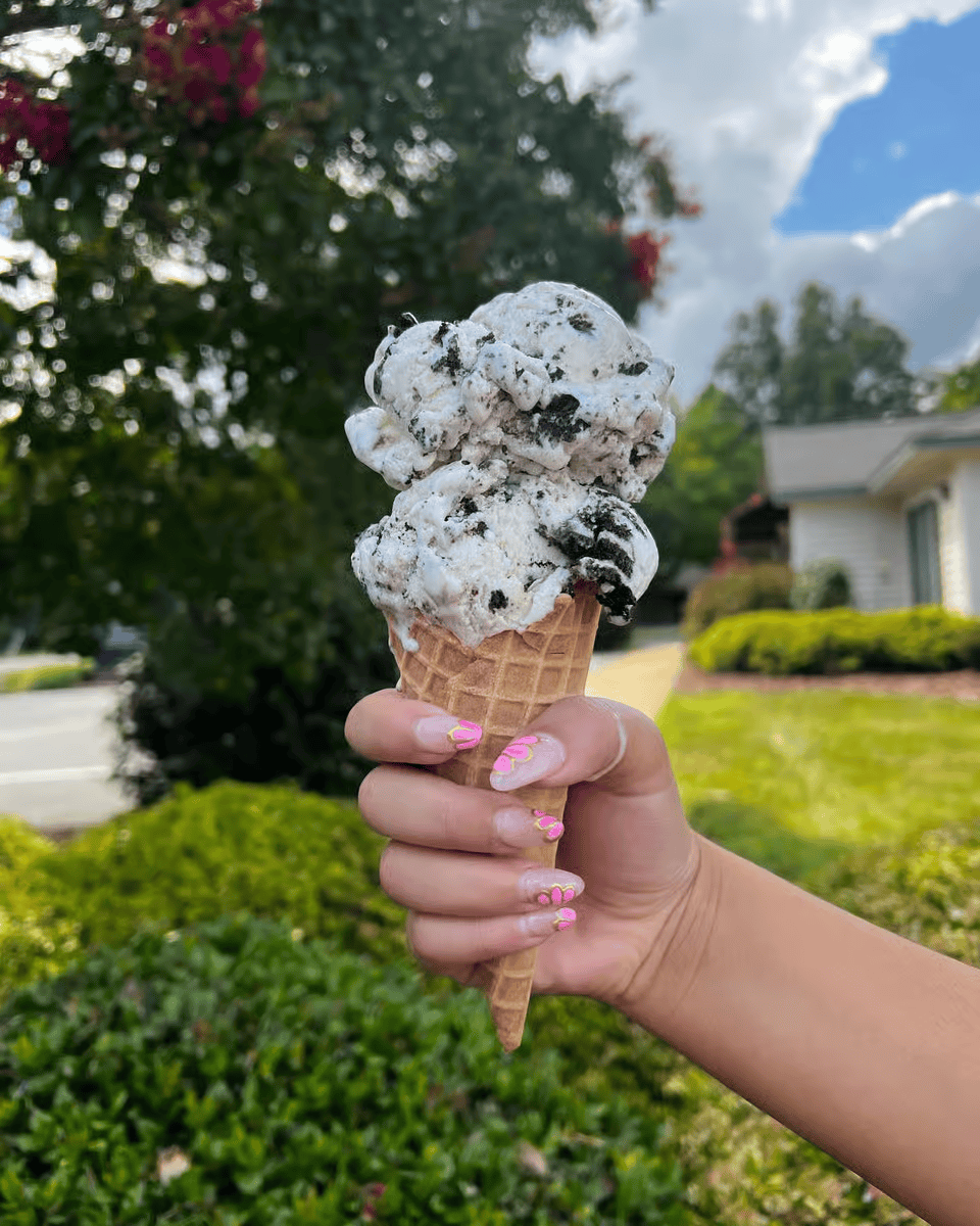 Hand holding a black-and-white ice cream cone outdoors near greenery and houses