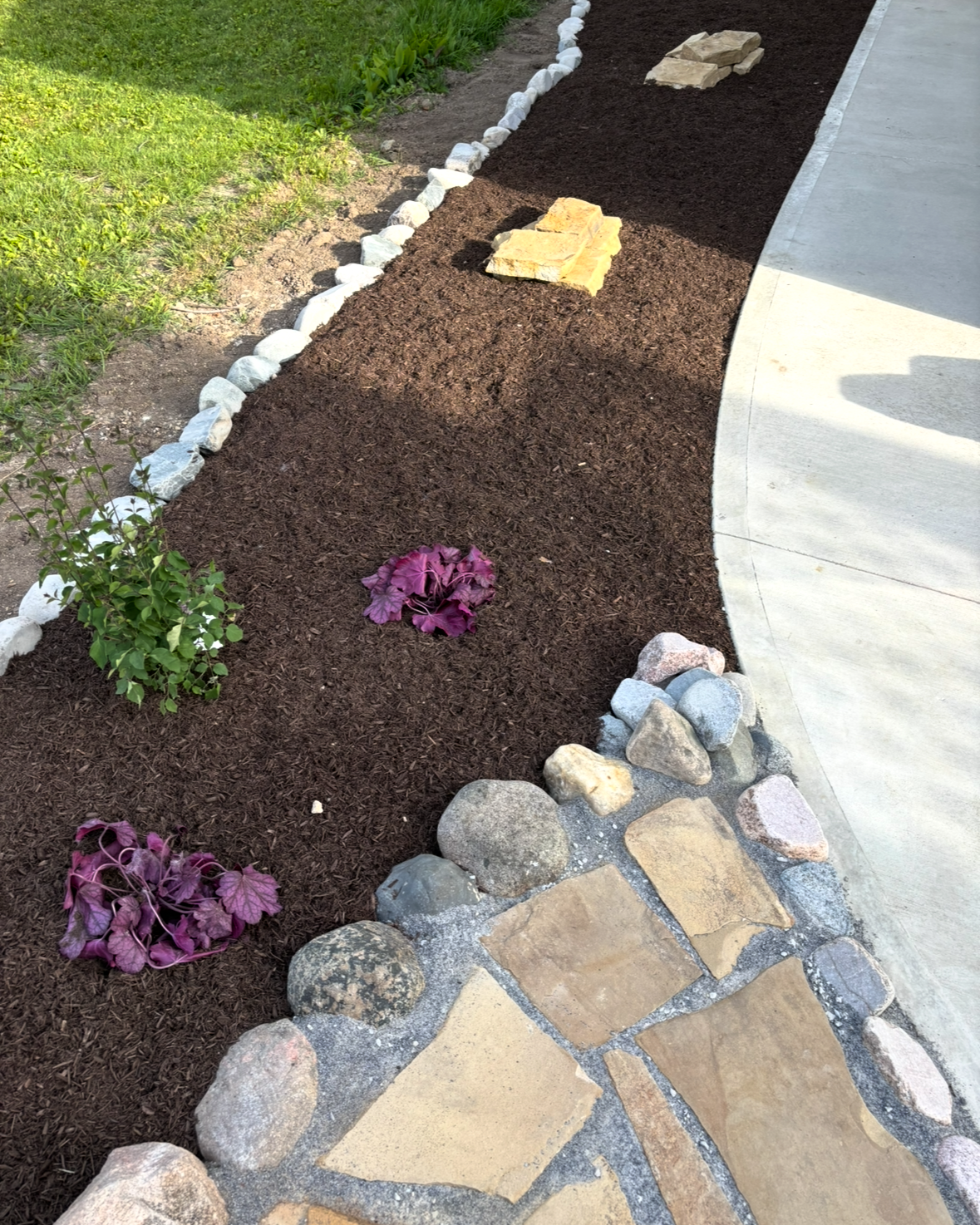 Curved garden bed with mulch, small plants, stone border, and a walkway edge