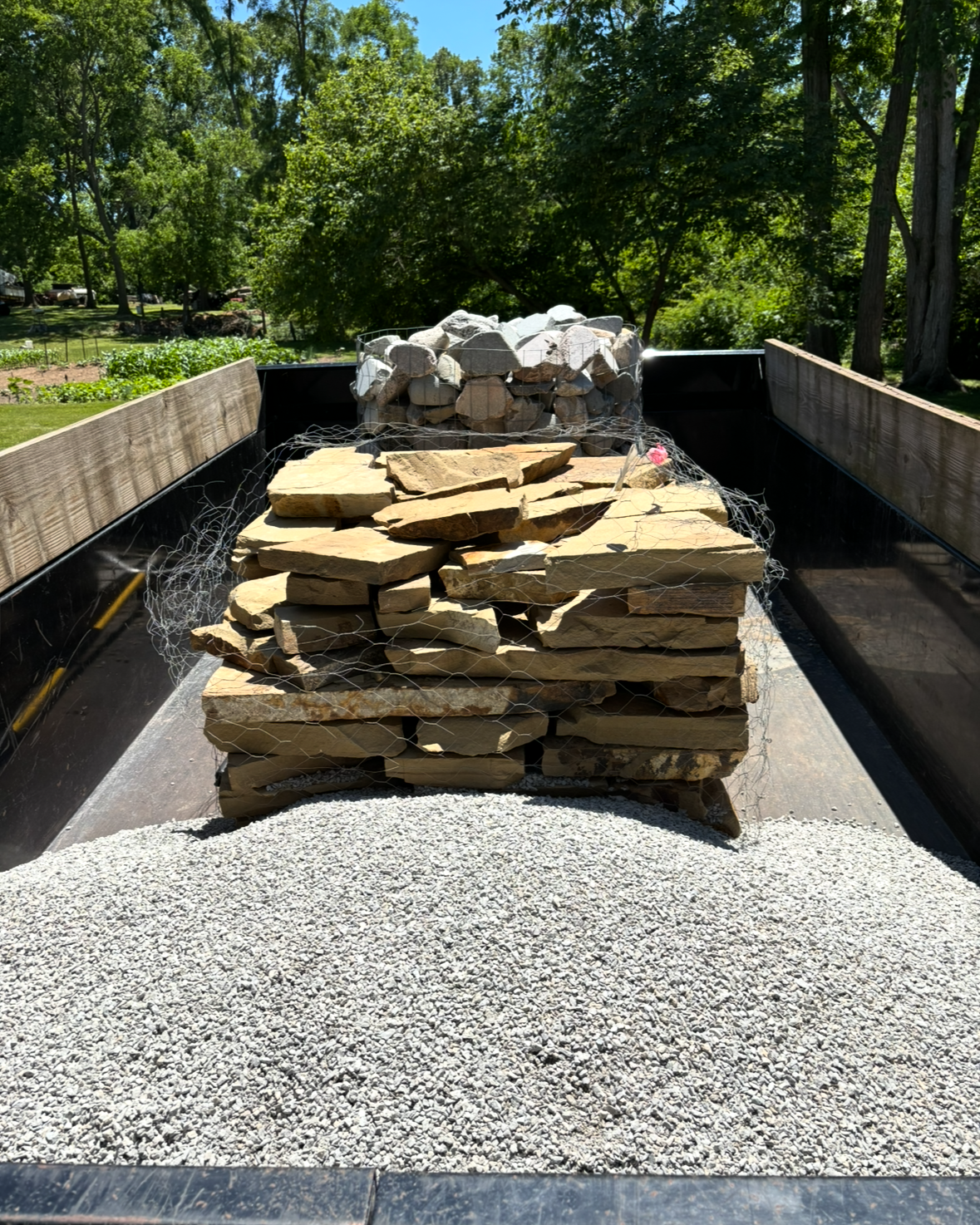 Stacked stone slabs in a narrow outdoor truck bed, with gravel and trees in the background.