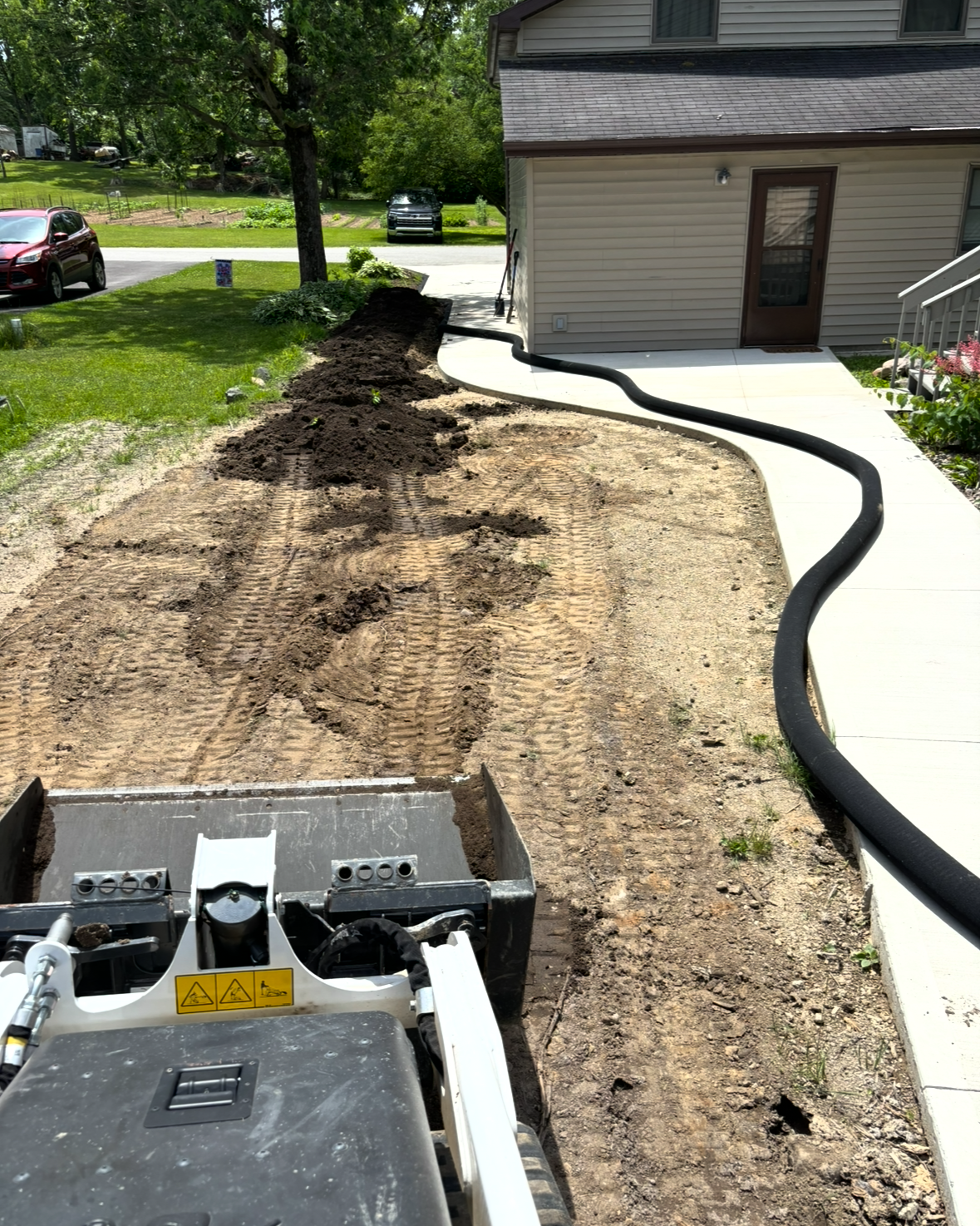 Backyard path work beside a house, with torn-up dirt and black drainage pipe along a concrete walkway.