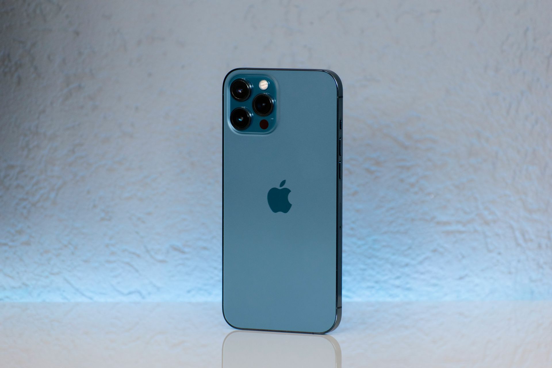 Blue iPhone standing upright on a white surface against a light gray wall with blue accent lighting