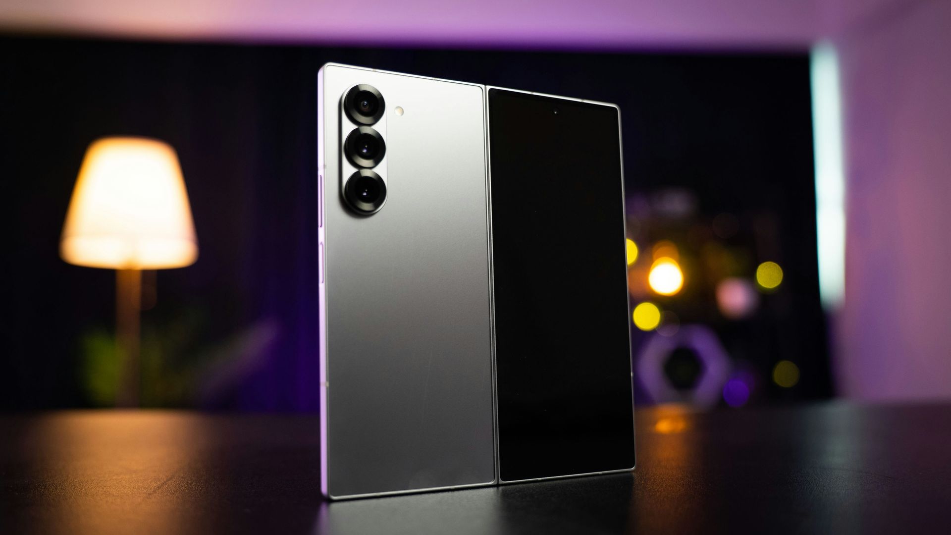 Silver foldable smartphone standing on a table with blurred lamp and lights in the background