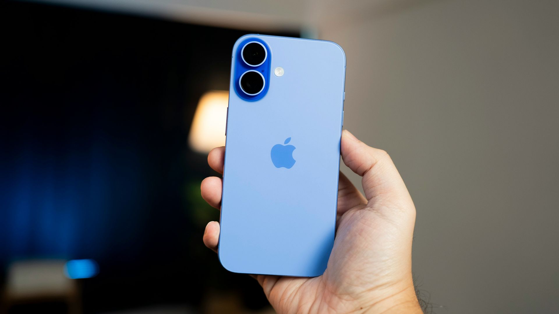 A hand holding a light blue iPhone with dual cameras against a blurred indoor background