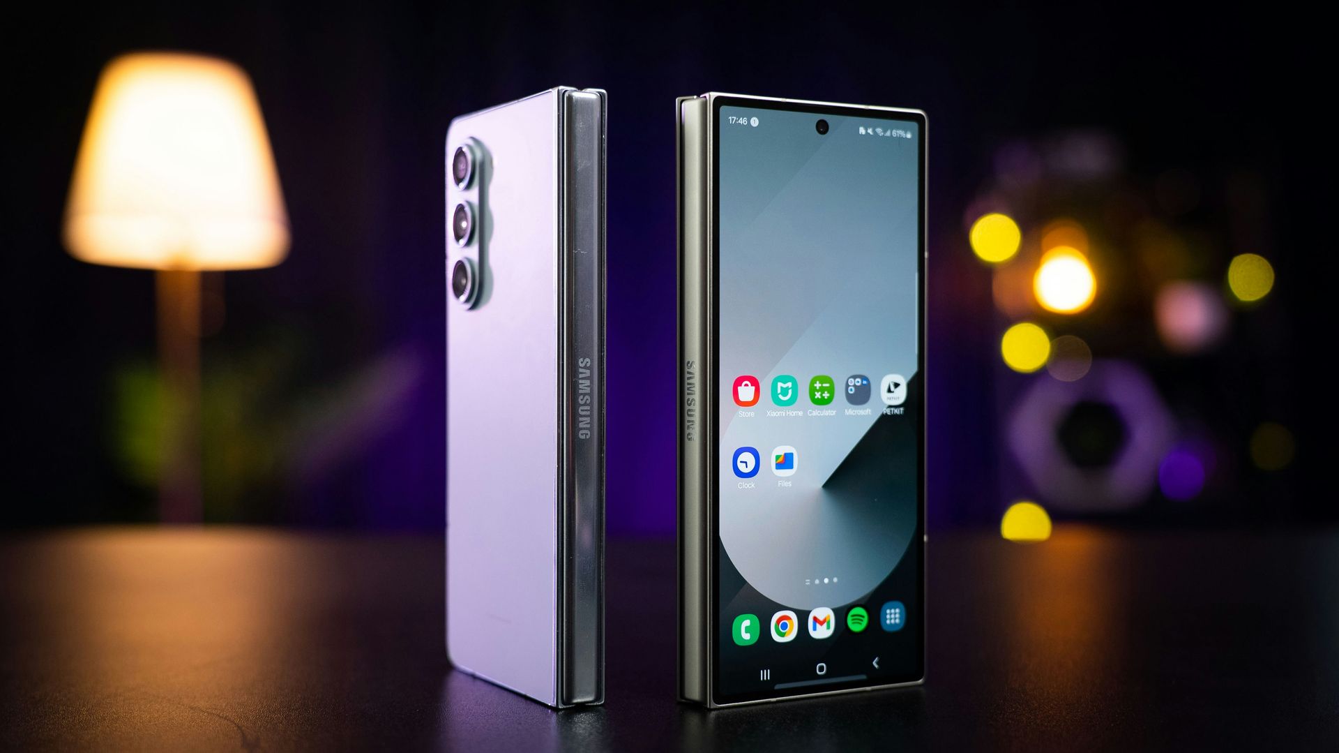 Foldable smartphone standing open on a table, showing front and back with a blurred lamp-lit background.
