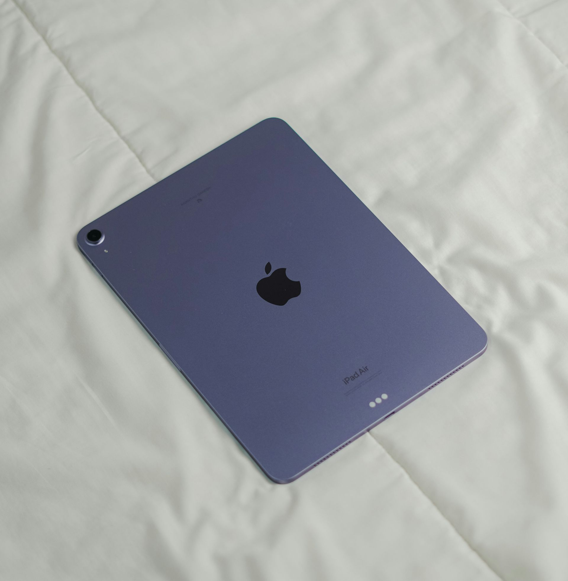 Purple iPad lying on a white bedspread, back side visible with Apple logo.