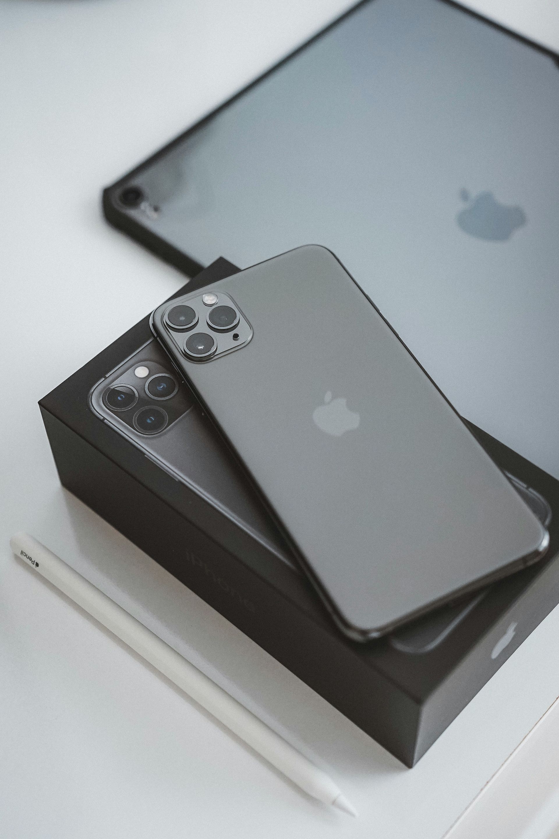 Gray iPhone with triple cameras resting on a black box, with a silver iPad in the background