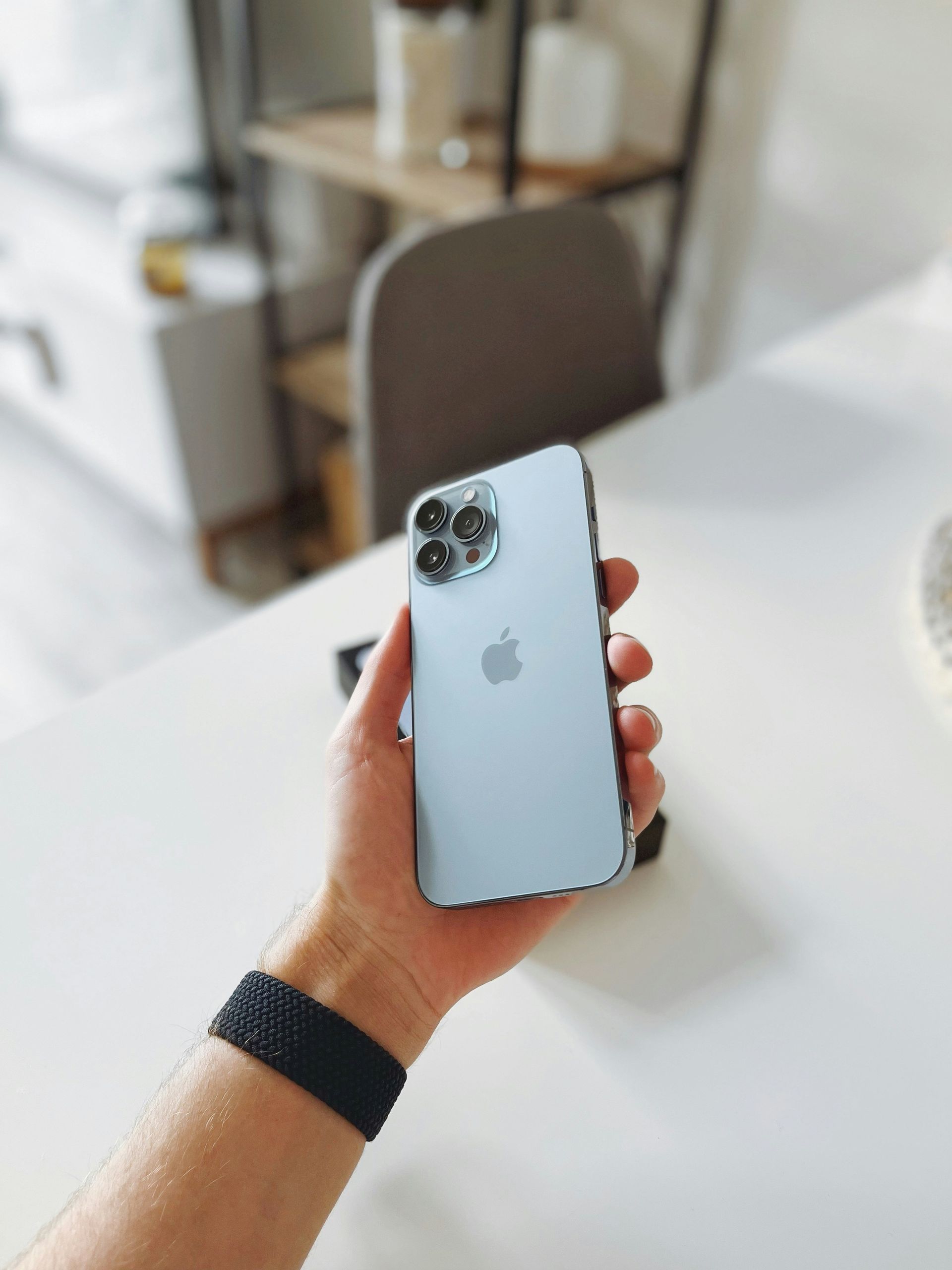 Hand holding a light blue iPhone over a white desk, with a blurred chair and shelving in the background.