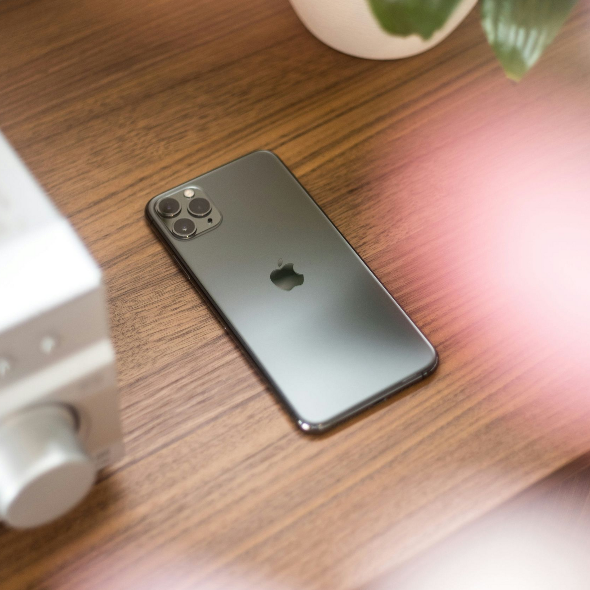 Silver iPhone lying on a wooden desk next to a white device, with a plant in the background.