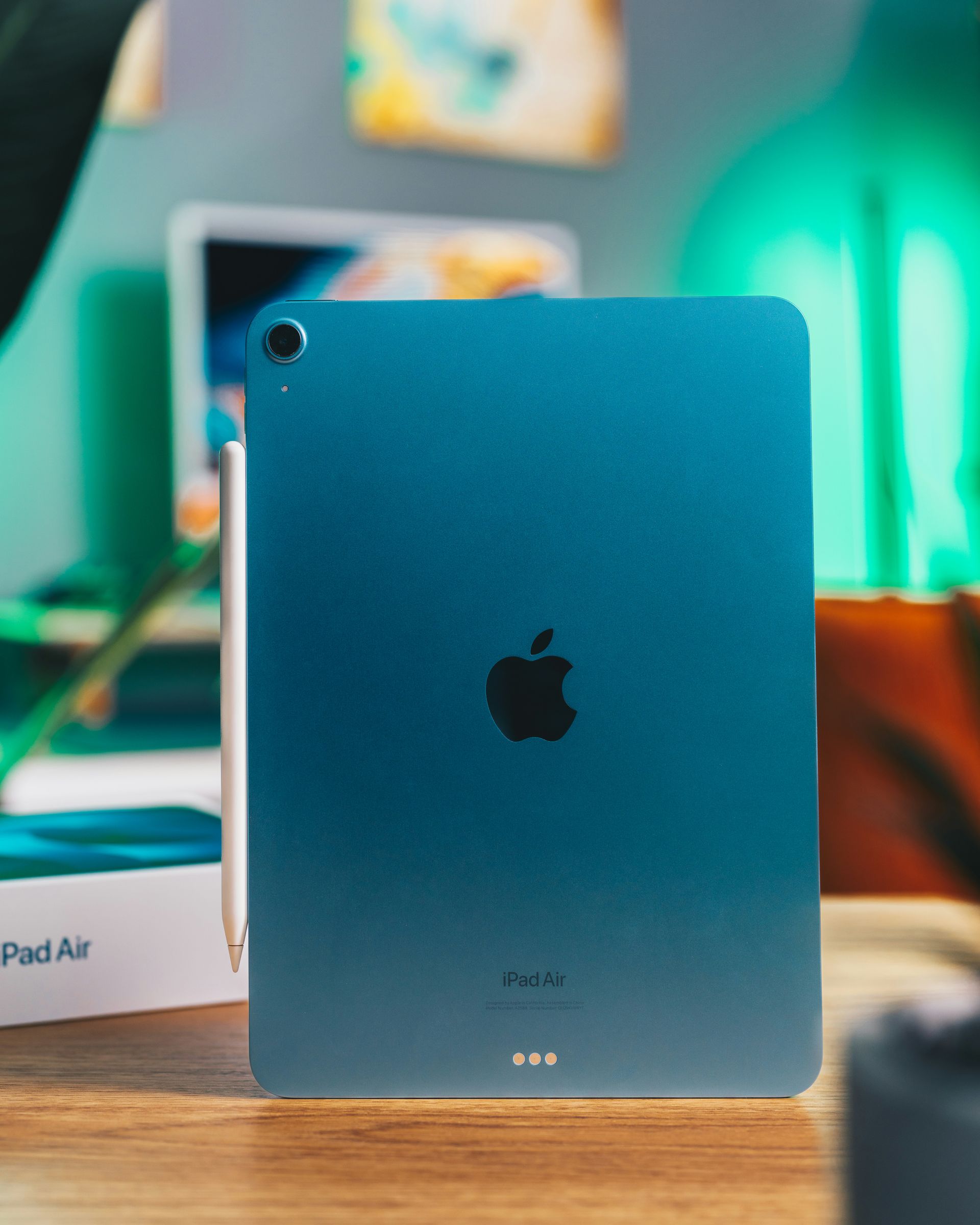 Blue iPad Air standing upright on a desk, with Apple logo visible and a blurred laptop in the background