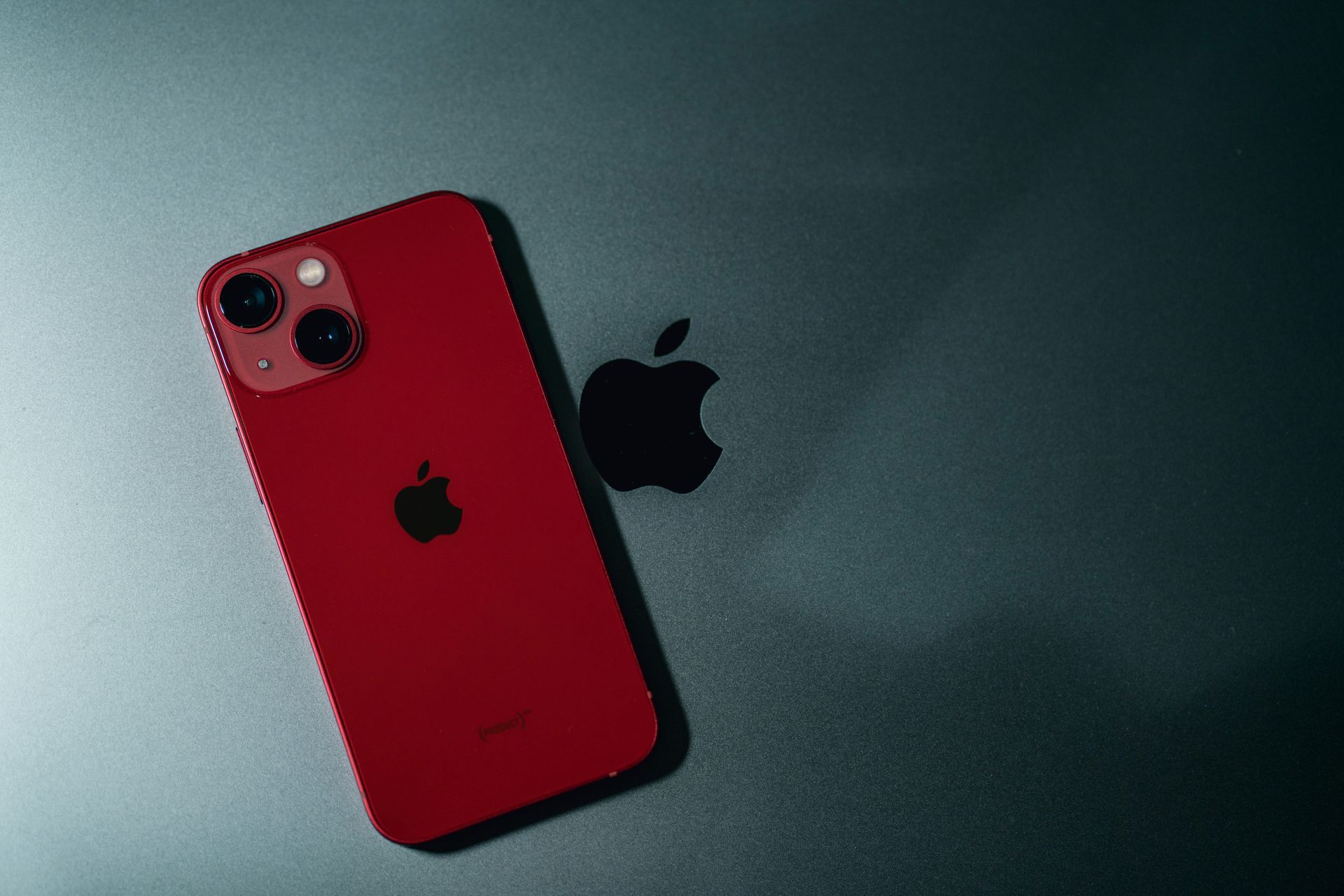 Red iPhone on a dark surface next to a black Apple logo.