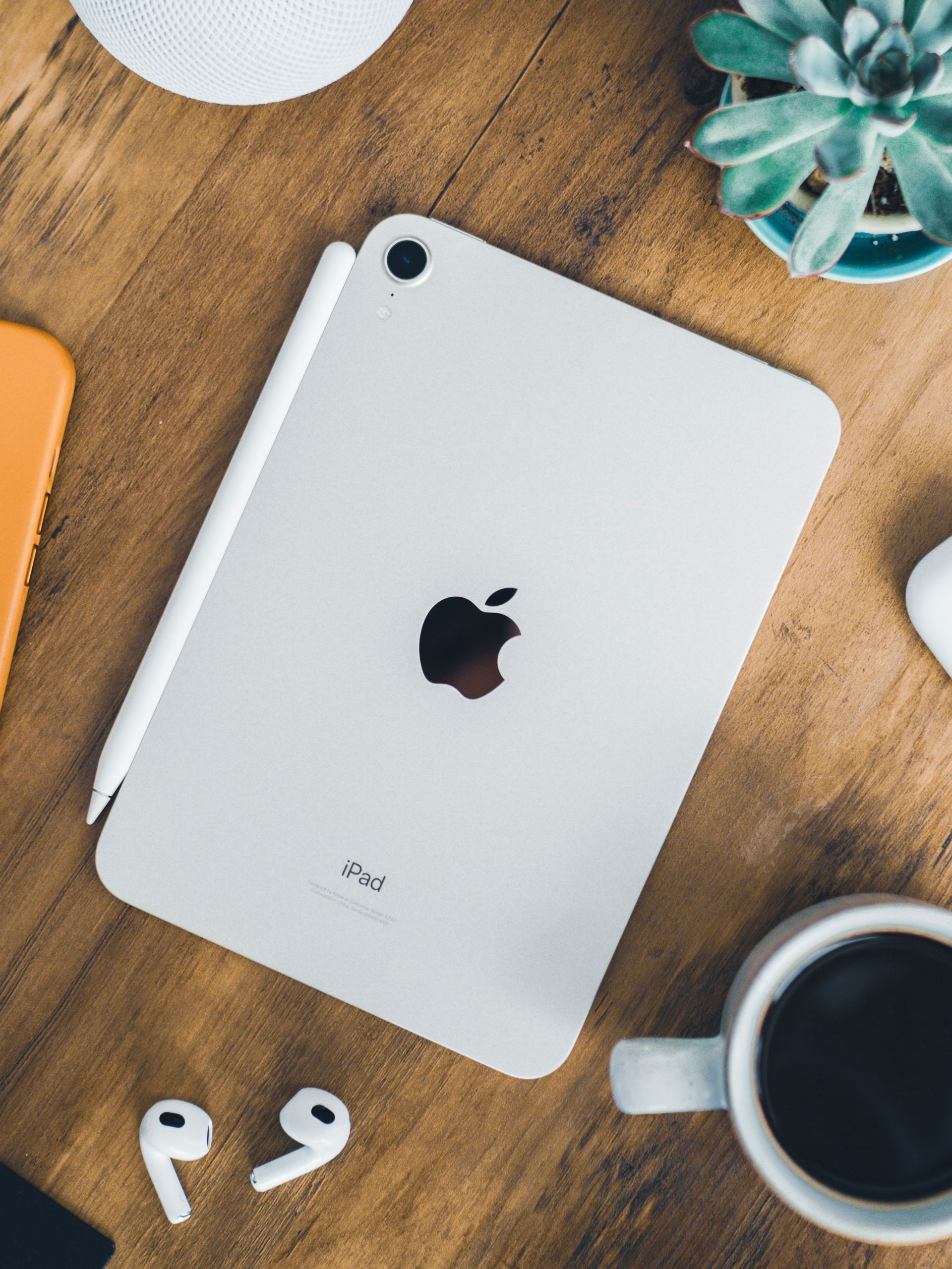 Silver iPad on a wooden desk with a coffee cup, plant, and earbuds nearby