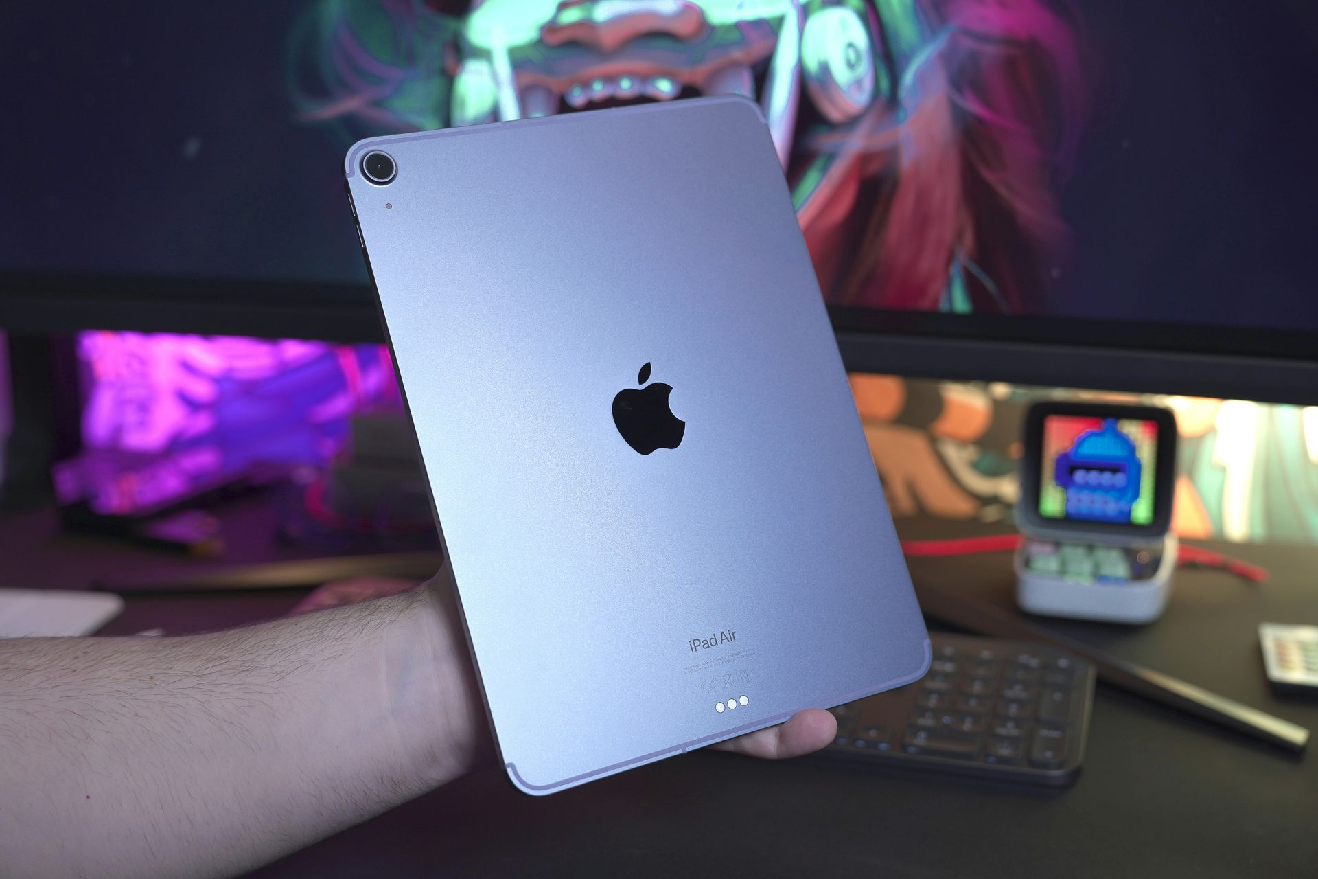 Silver iPad held in front of a colorful desk setup with a monitor and small keyboard in the background