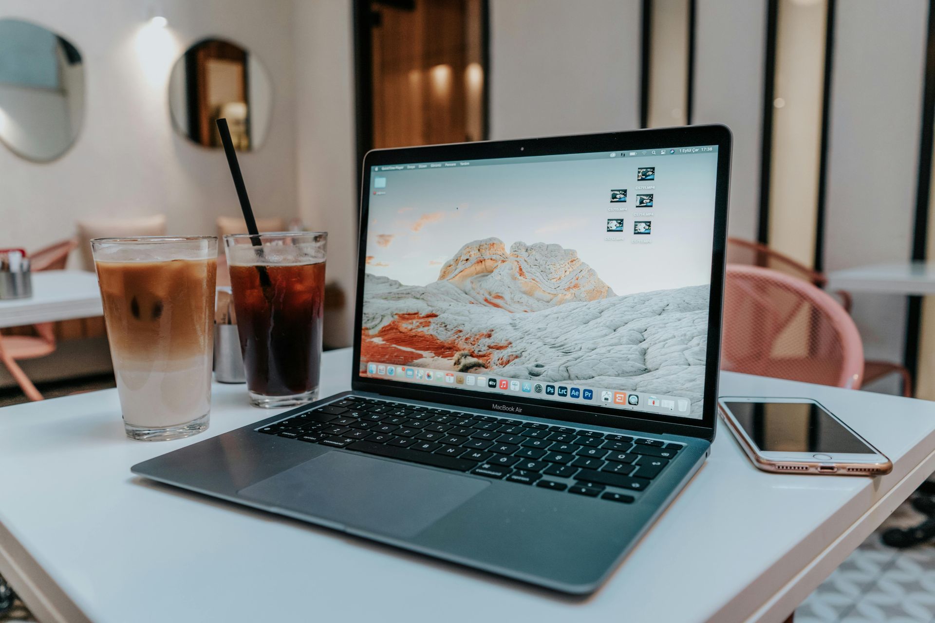 Laptop on a cafe table beside iced drinks and a smartphone, with a mountain wallpaper on screen