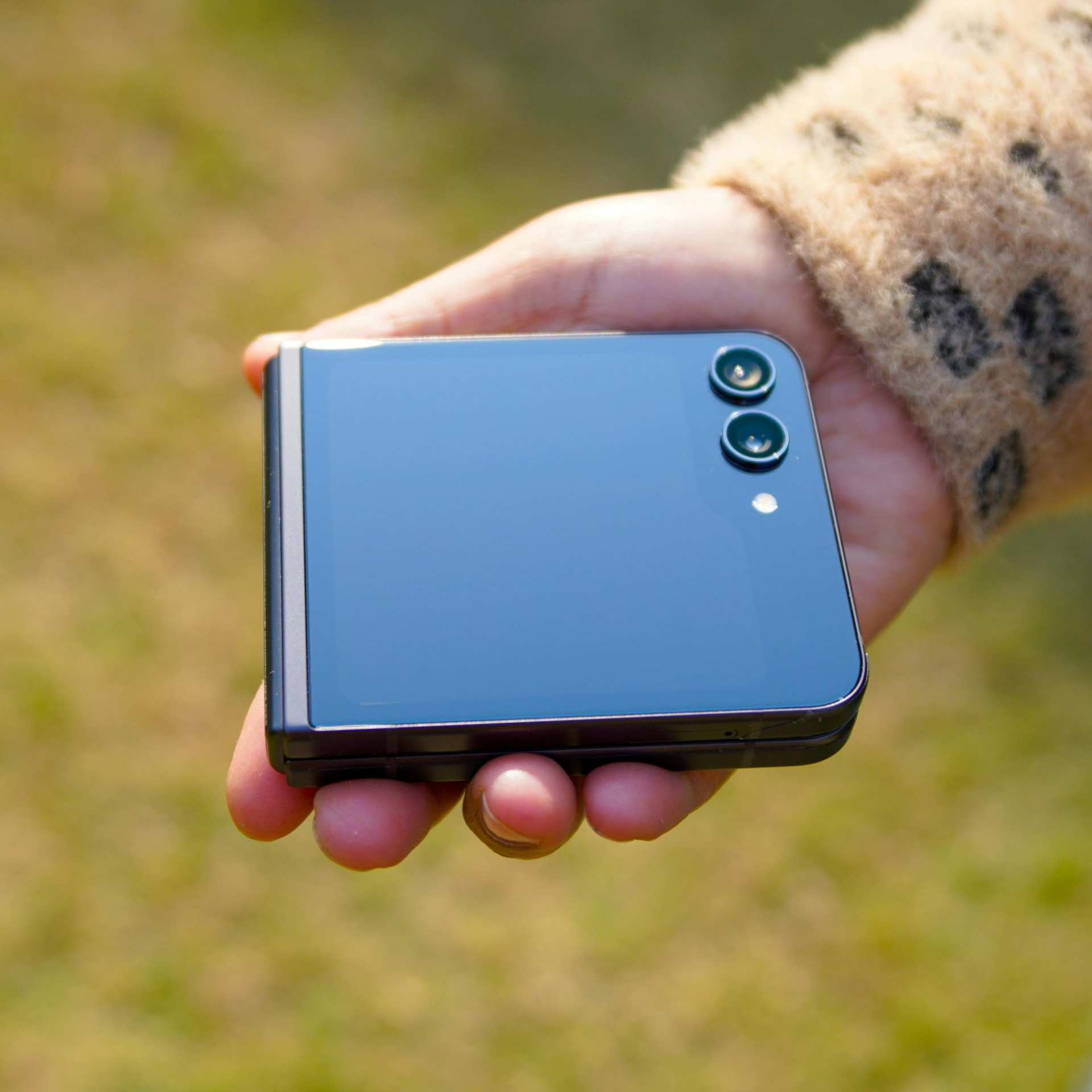 Hand holding a closed glossy blue folding smartphone outdoors on grass