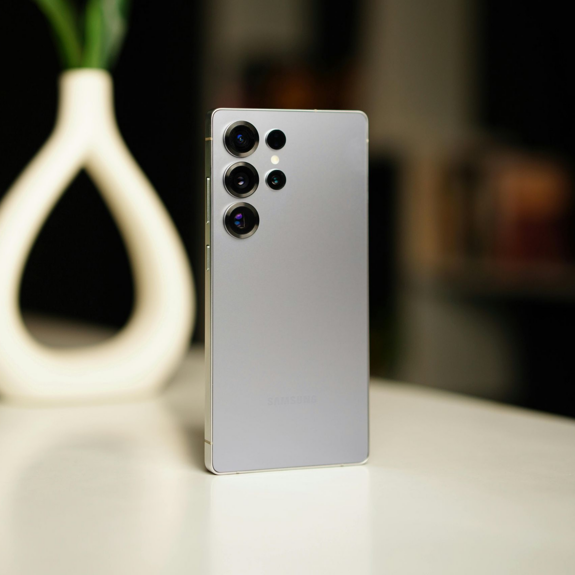 Silver smartphone standing upright on a table, with three rear cameras and a blurred lamp in the background