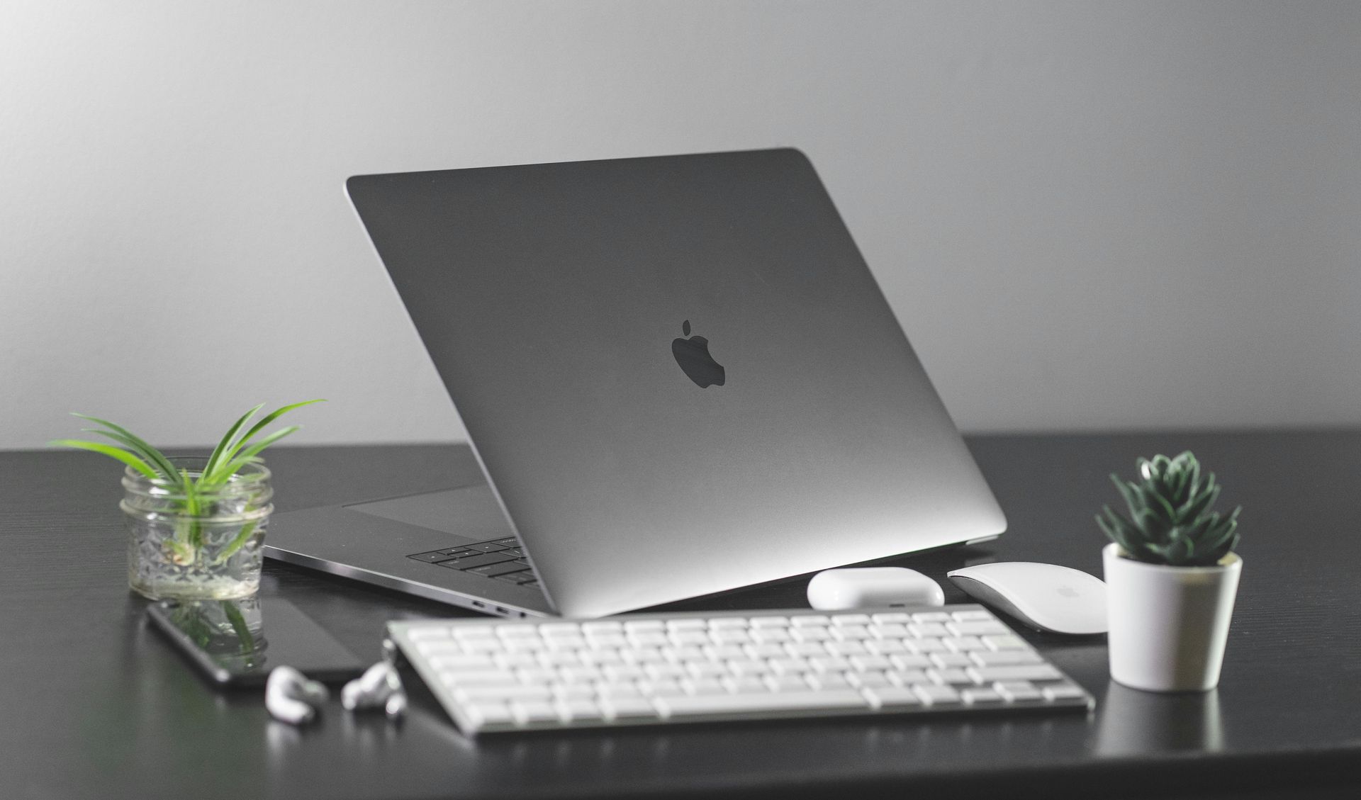 MacBook on desk with white keyboard, mouse, earbuds, and small plants against a gray wall
