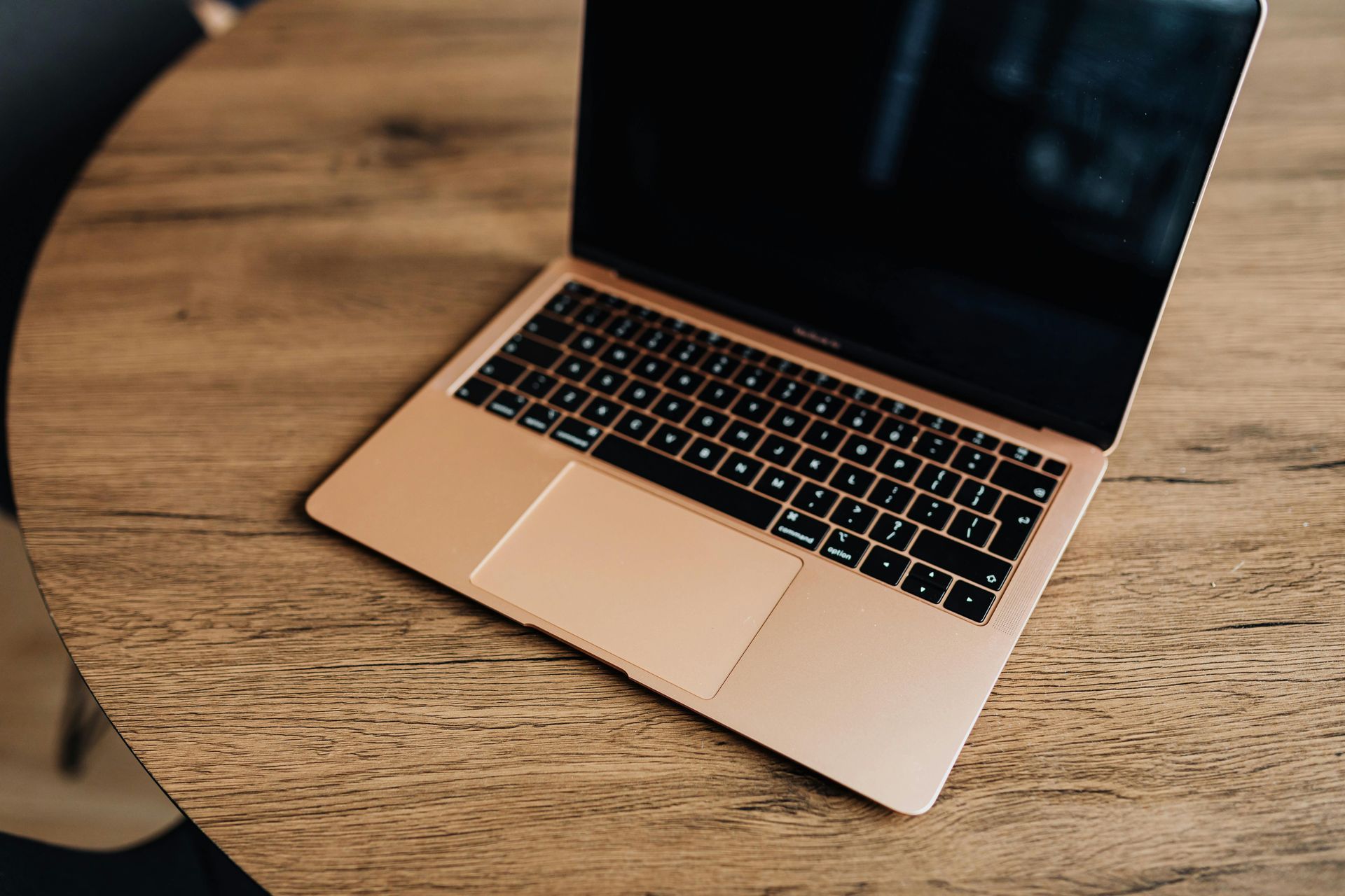 Open laptop on a wooden table, showing a black screen and rose-gold keyboard.