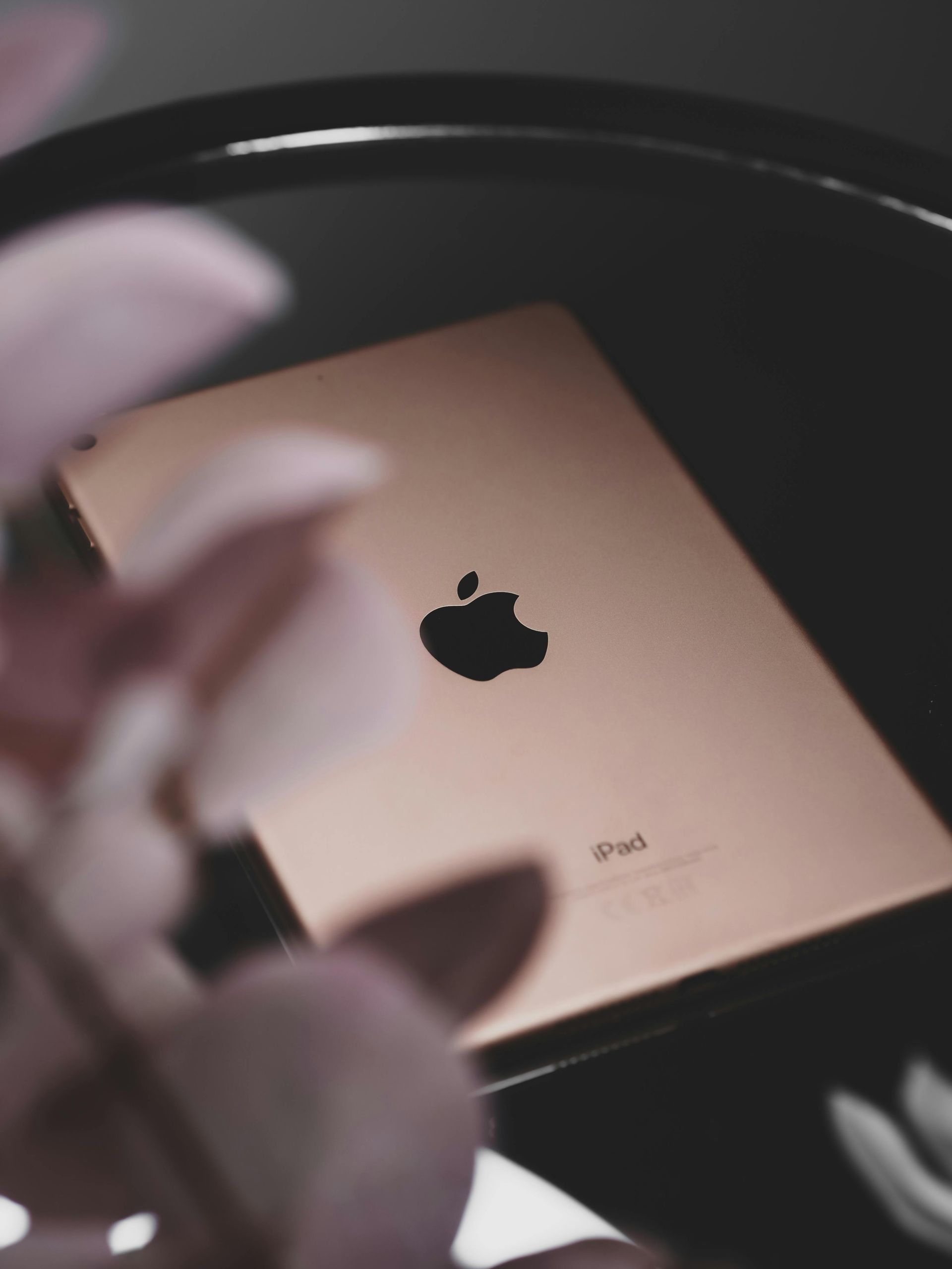 Rose gold iPhone with Apple logo on a dark surface, partially framed by a blurred hand and flowers