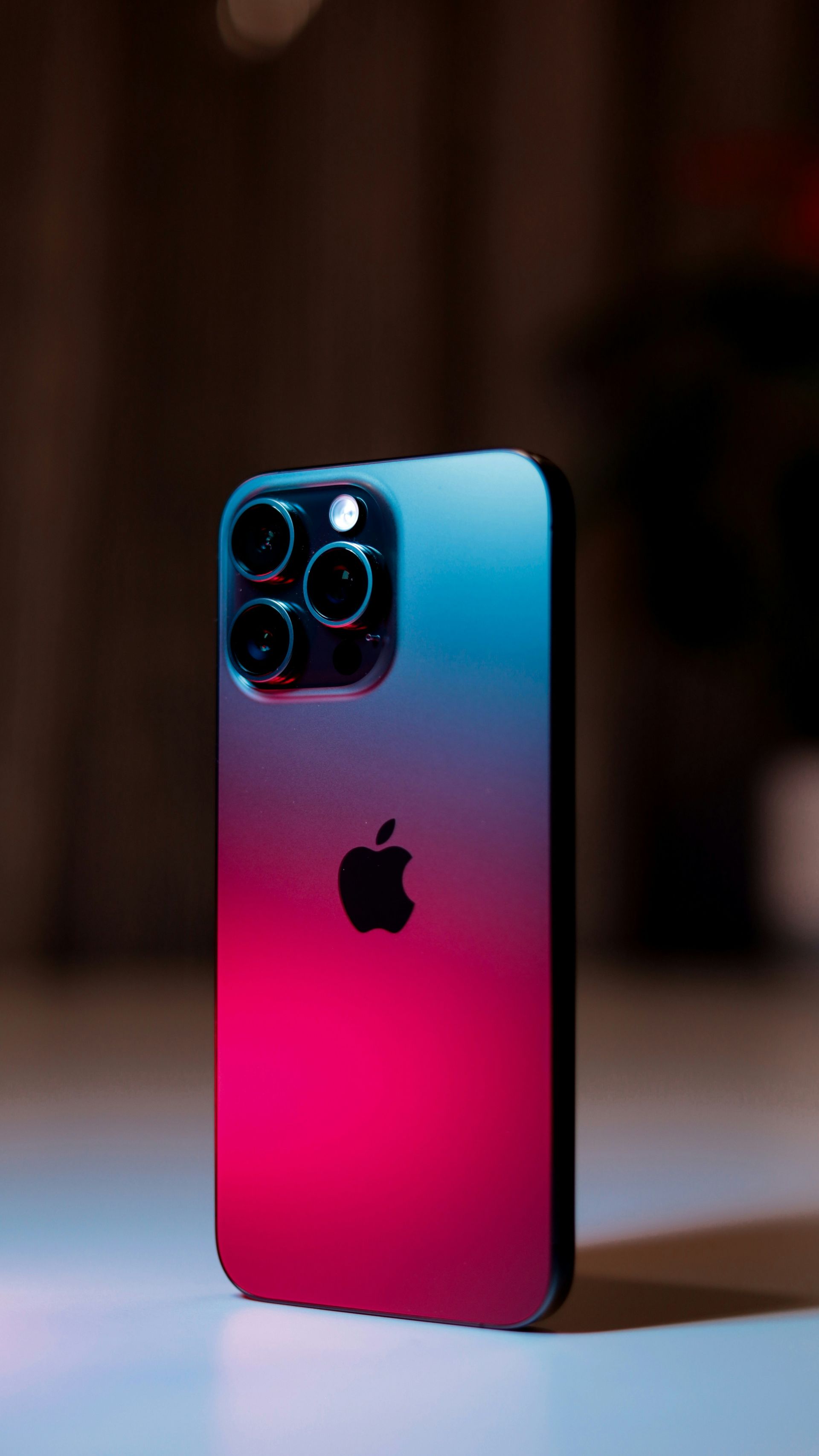 Apple iPhone with triple camera, blue-to-pink gradient back, standing upright on a surface