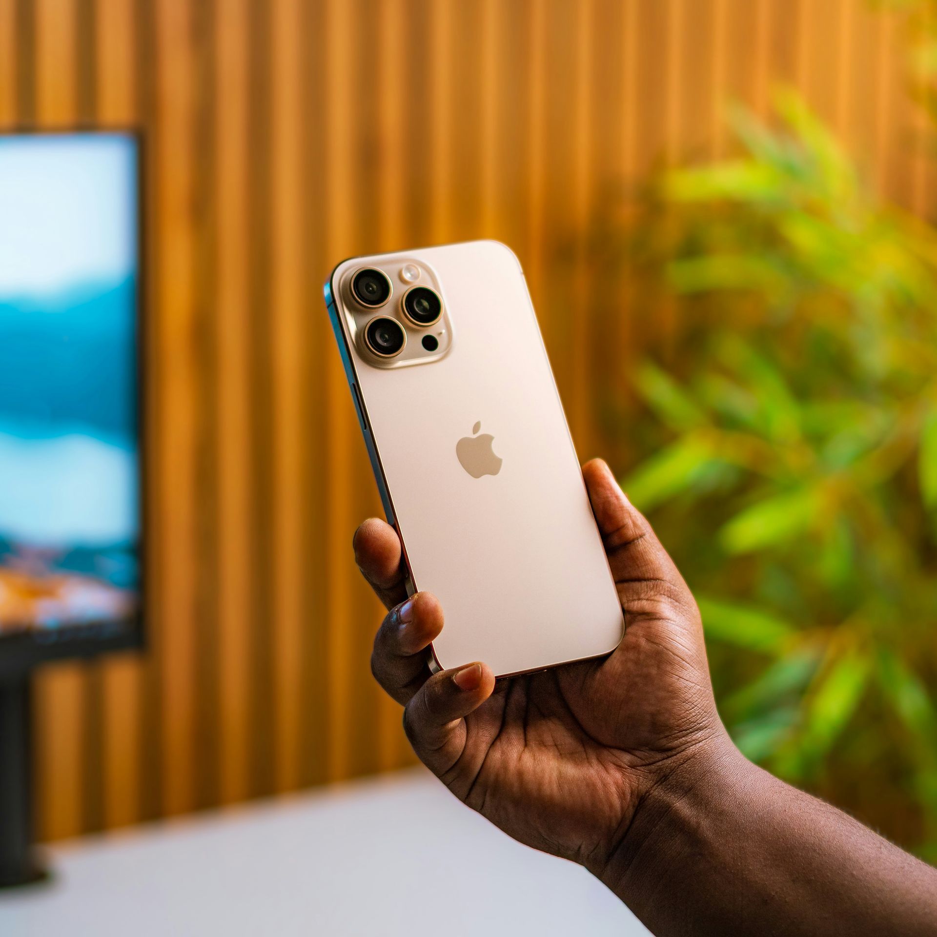 Hand holding a gold iPhone with triple camera, against a blurred indoor background