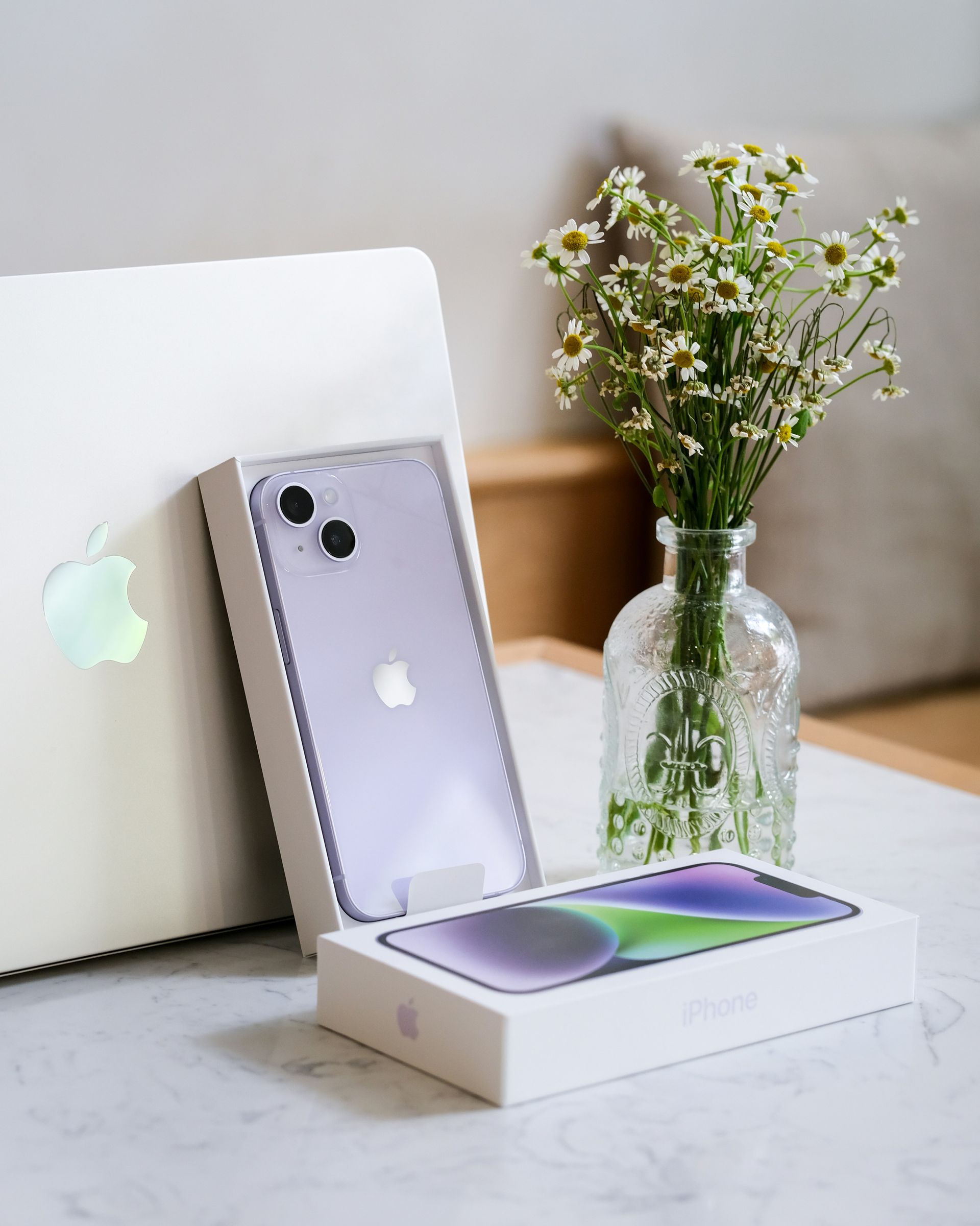 IPhone, MacBook, and flowers on a white desk with a boxed phone in the foreground