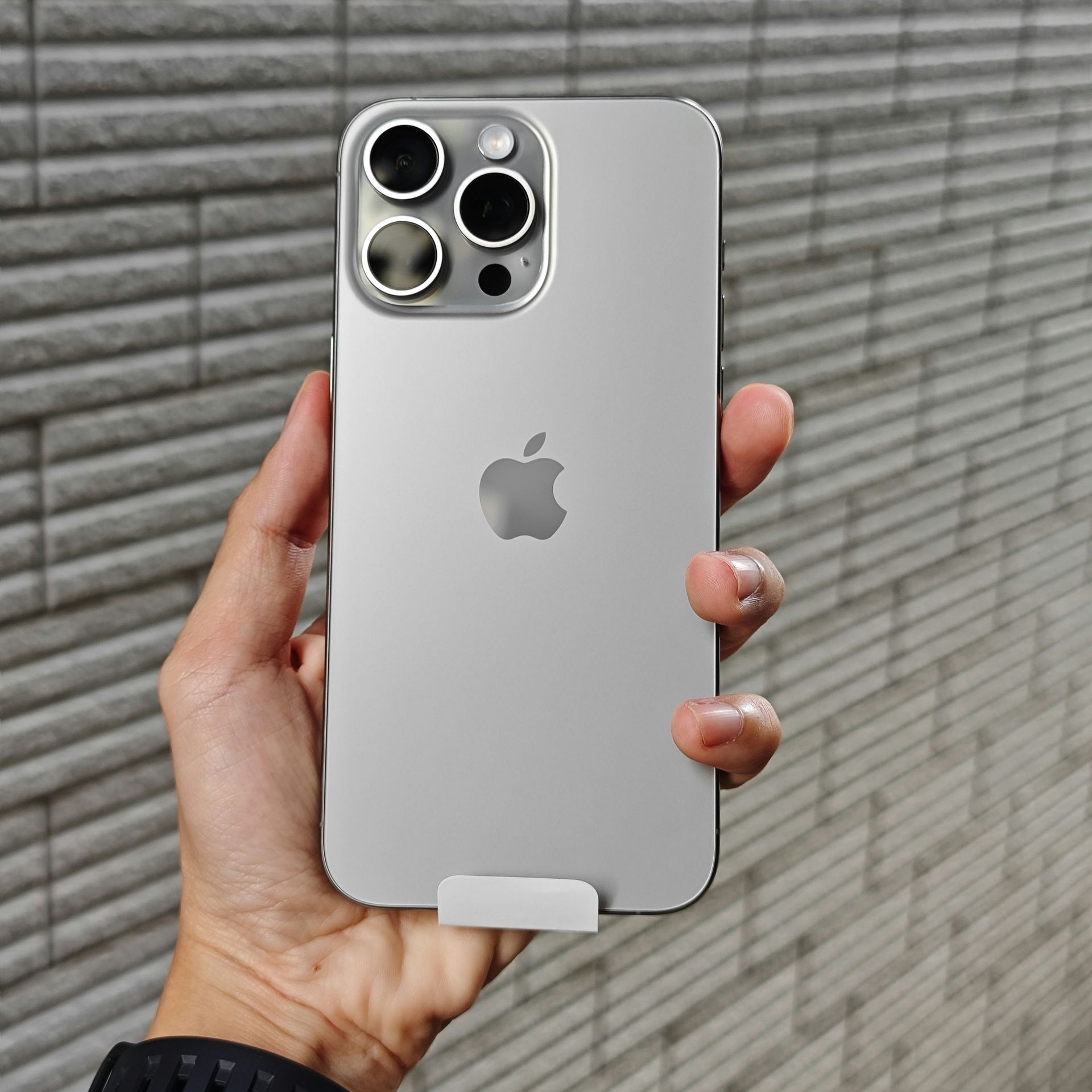 Hand holding a silver iPhone with triple rear cameras against a gray tiled wall