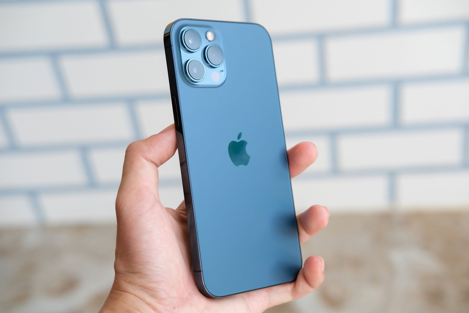 Hand holding a blue iPhone with triple cameras against a blurred brick wall background