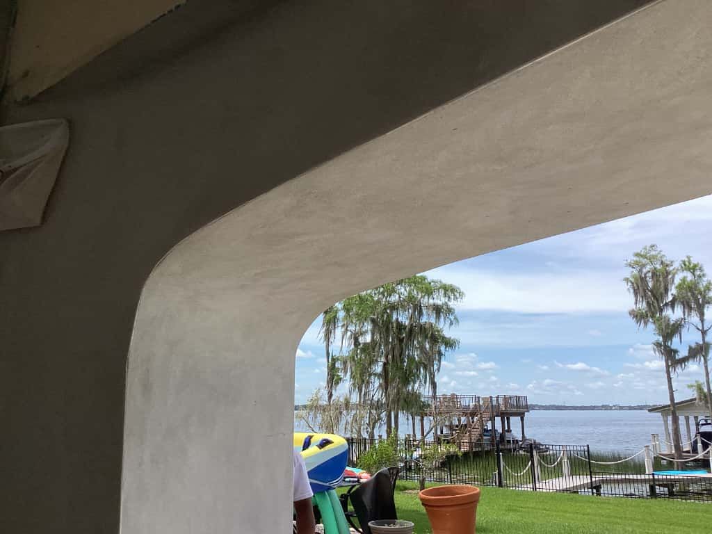 An arched doorway frames a view of a lake with trees, a dock, and a blue sky.