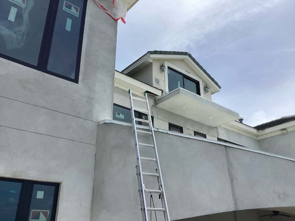 A tall ladder leans against a two-story house under construction, with gray stucco walls and dark window frames.