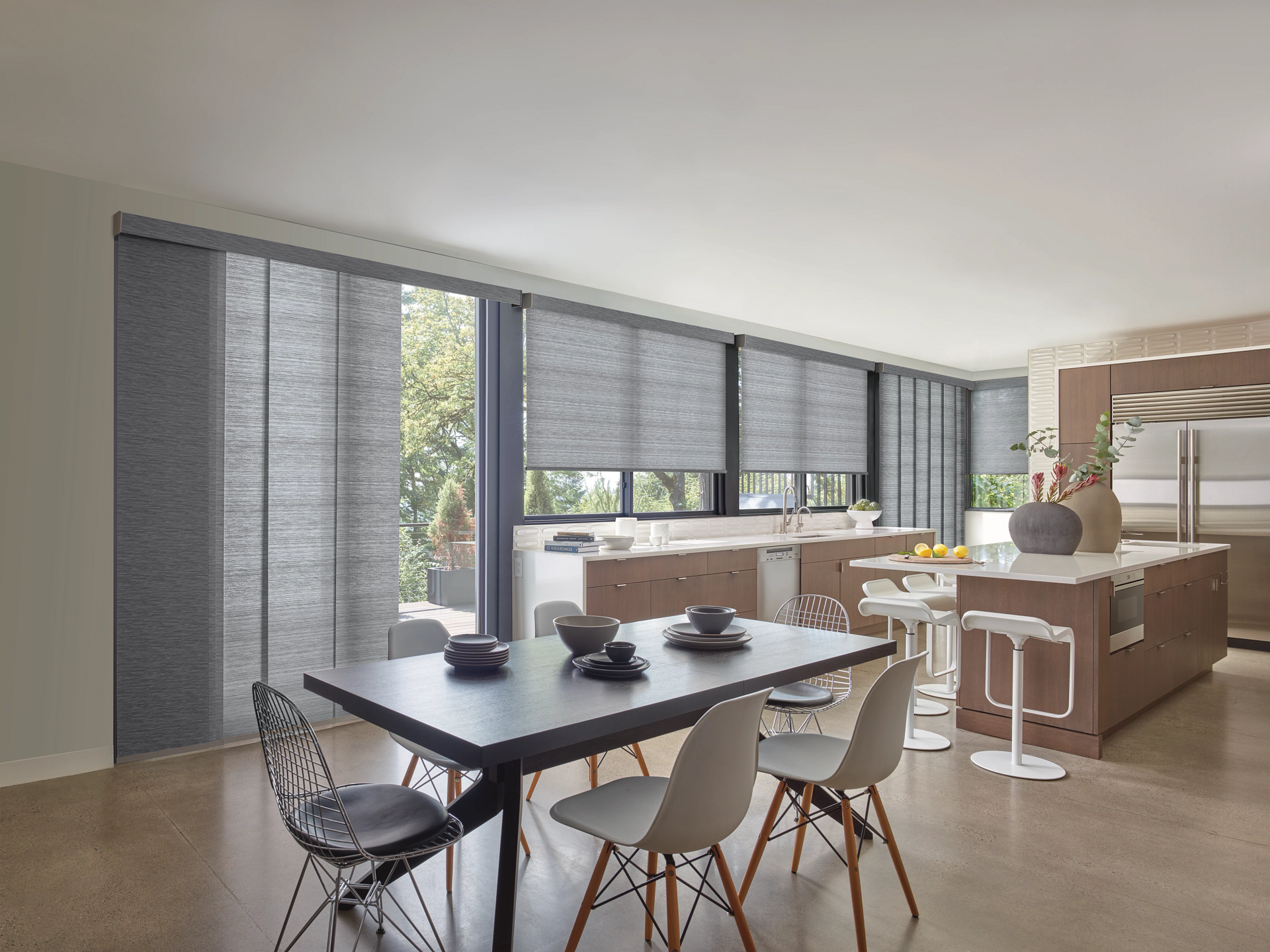Dining room with a black table, white chairs, and gray panel blinds overlooking a kitchen.