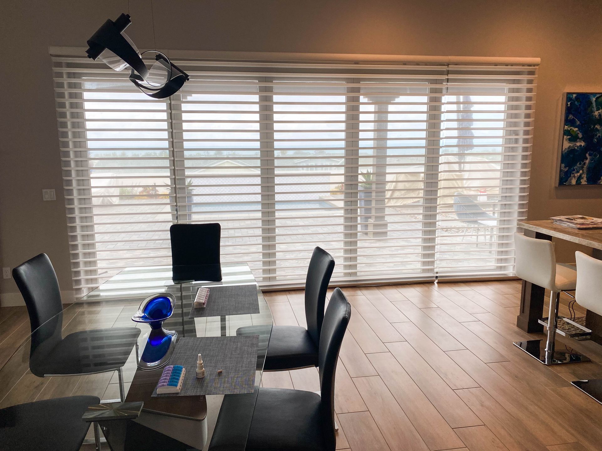 Dining room with table, chairs, large window with blinds, and blue artwork.