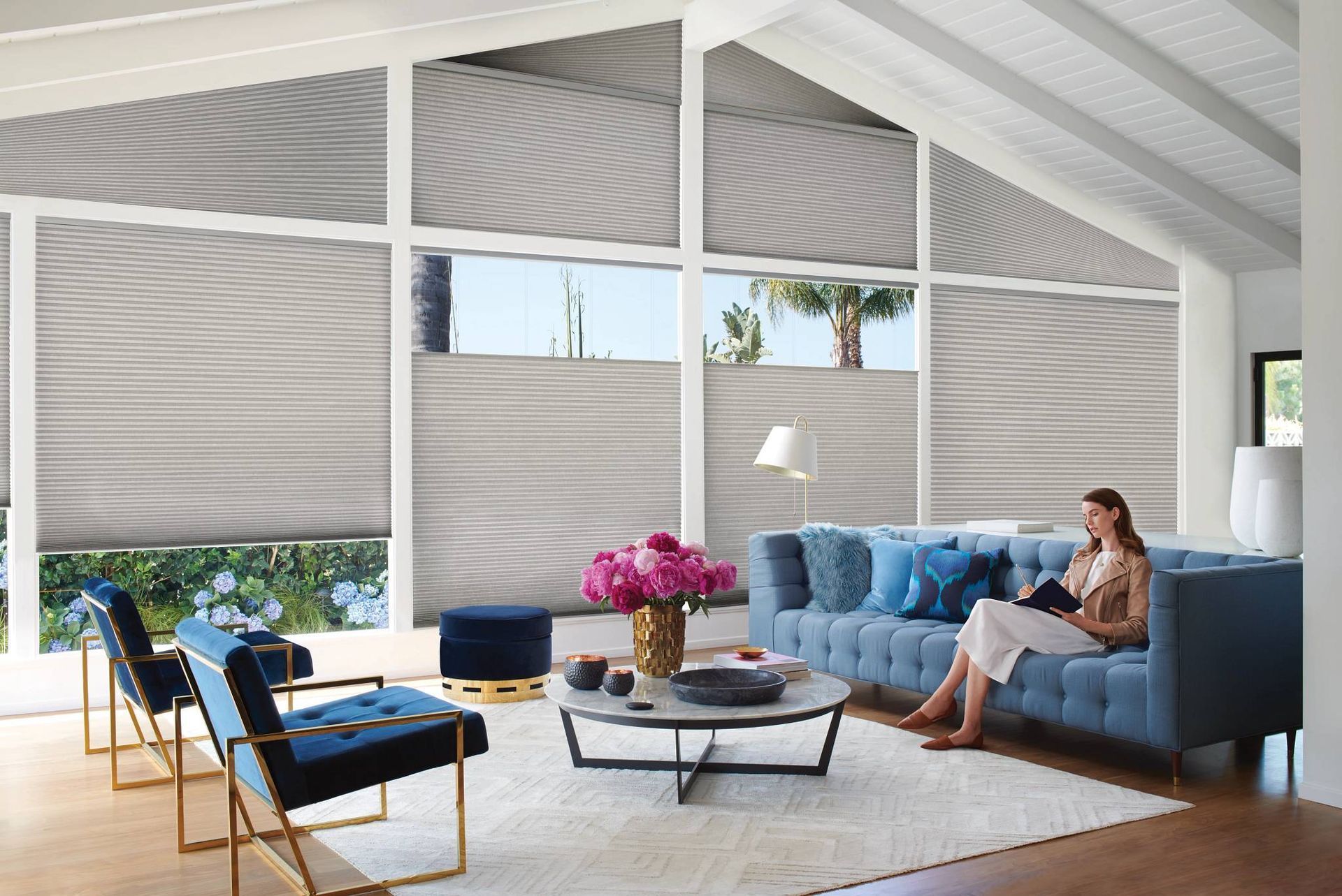 Living room with blue sofa, woman reading, large windows with shades.