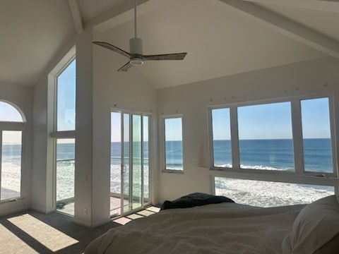 Bright bedroom with ocean view through large windows and balcony doors, white walls, and ceiling fan