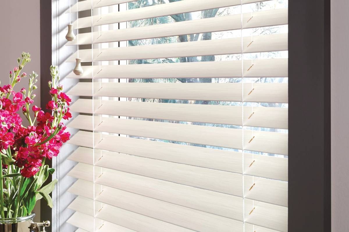 Window with closed, cream-colored blinds; vibrant pink flowers in a glass vase to the left.