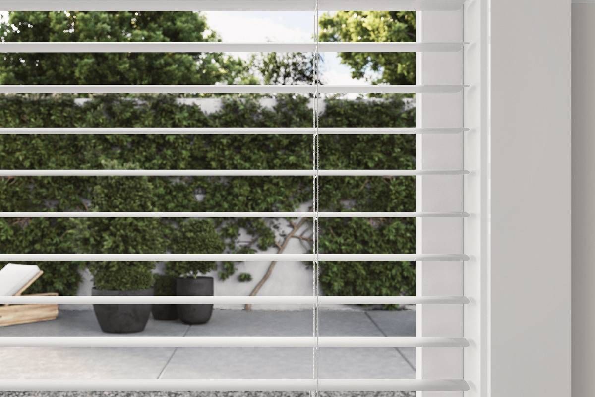 White blinds partially cover a view of a patio with plants and potted decor.