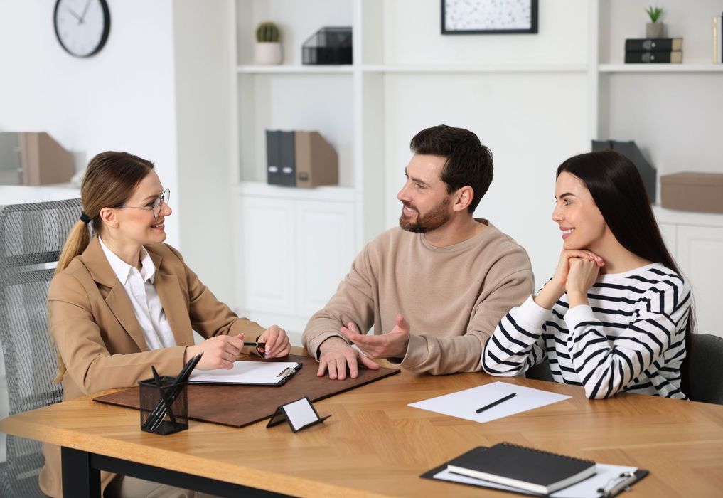 A professional in a tan blazer meets with a couple at a desk in a bright office, discussing documents during a meeting.