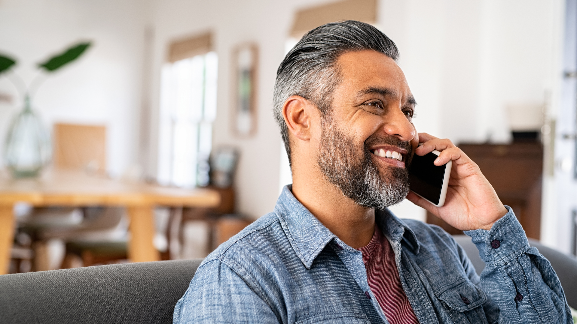 Smiling man on a couch talking on a smartphone in a bright living room